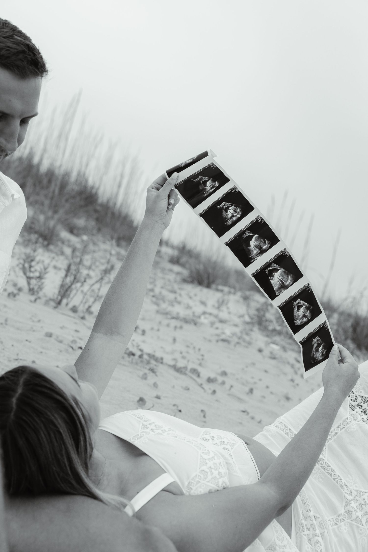 Black and white photo of couple looking at ultrasound images on a sandy beach.
