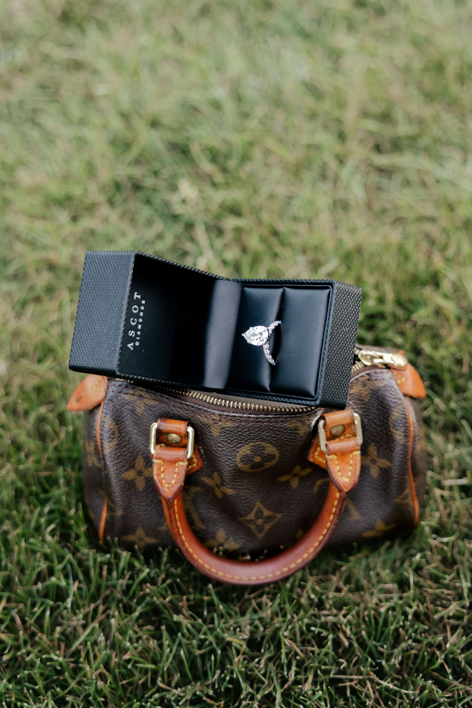 Diamond engagement ring in black Ascot jewelry box displayed on Louis Vuitton monogram mini bag on green grass.