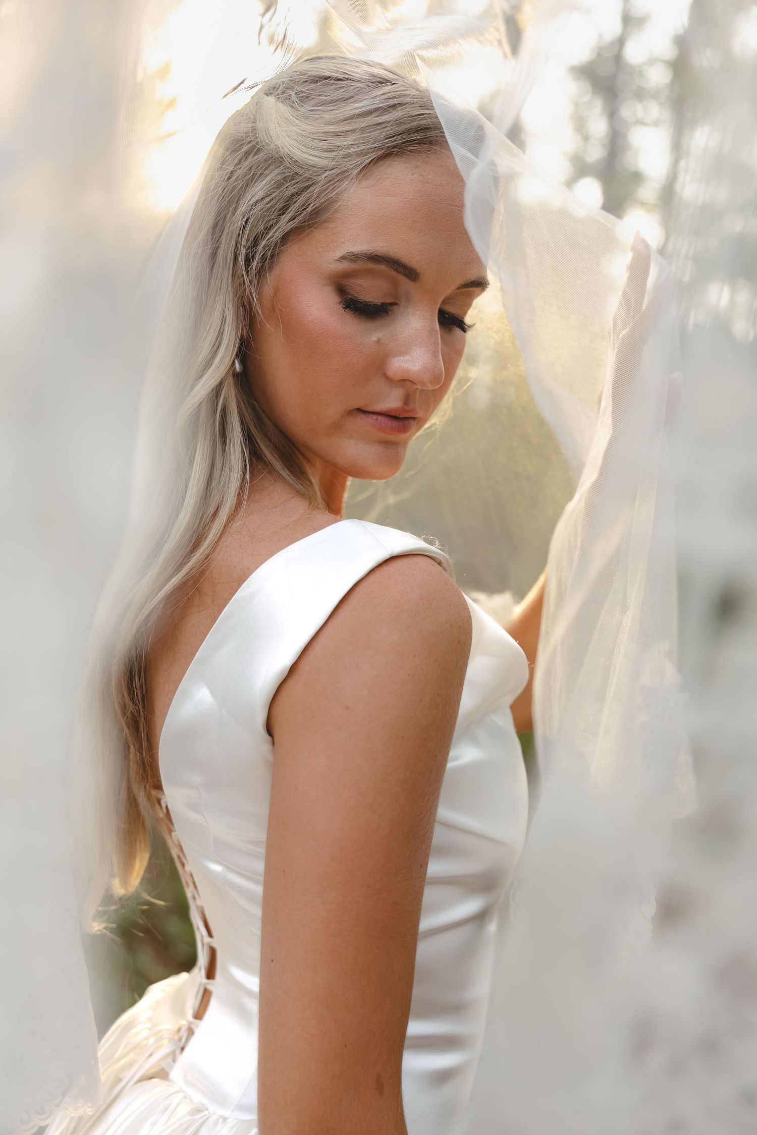 Blonde bride in white satin gown posing gracefully among sheer white veils.