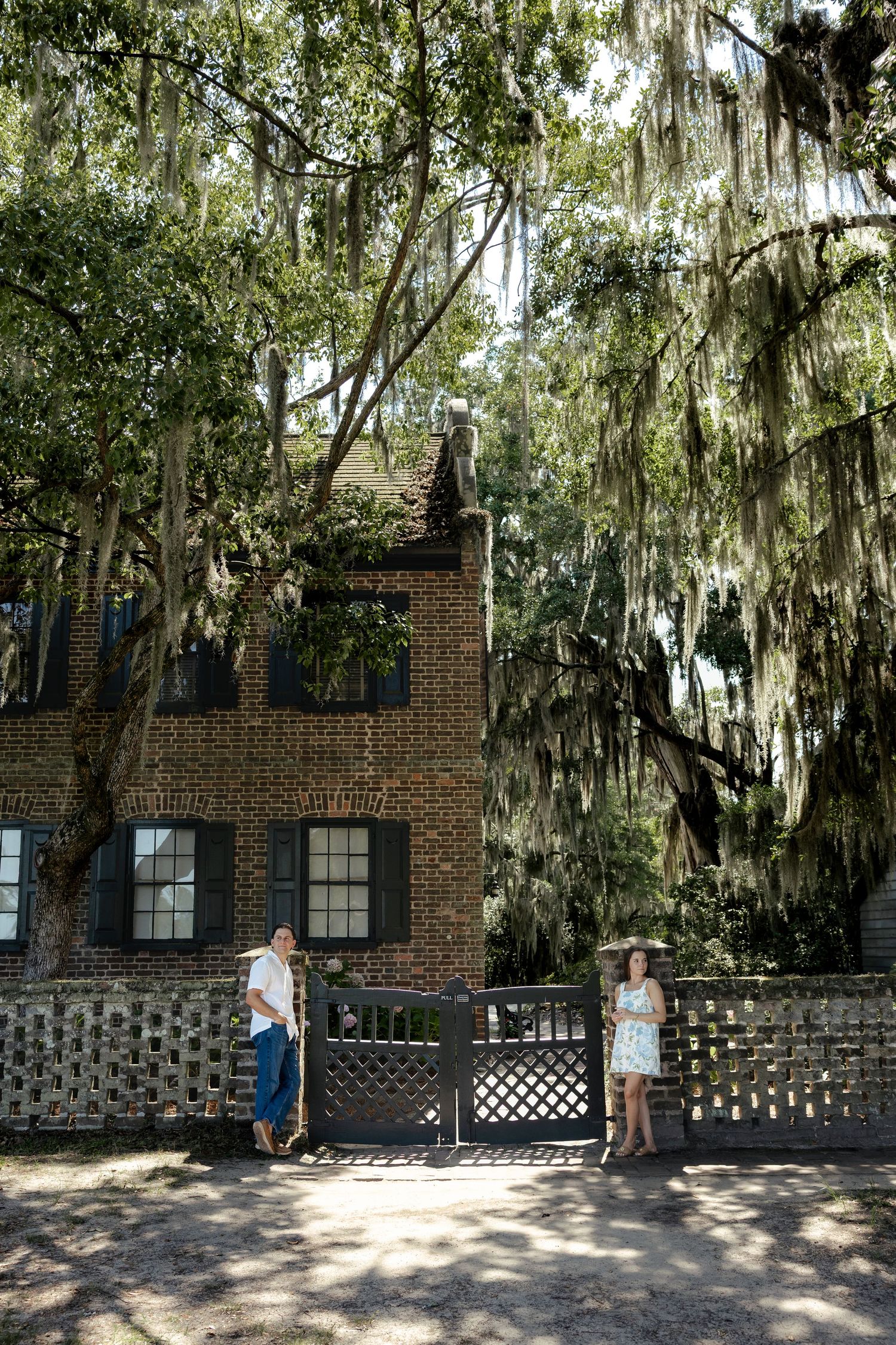 Couple standing at a garden gate in front of a historic brick building surrounded by Spanish moss-draped oak trees.