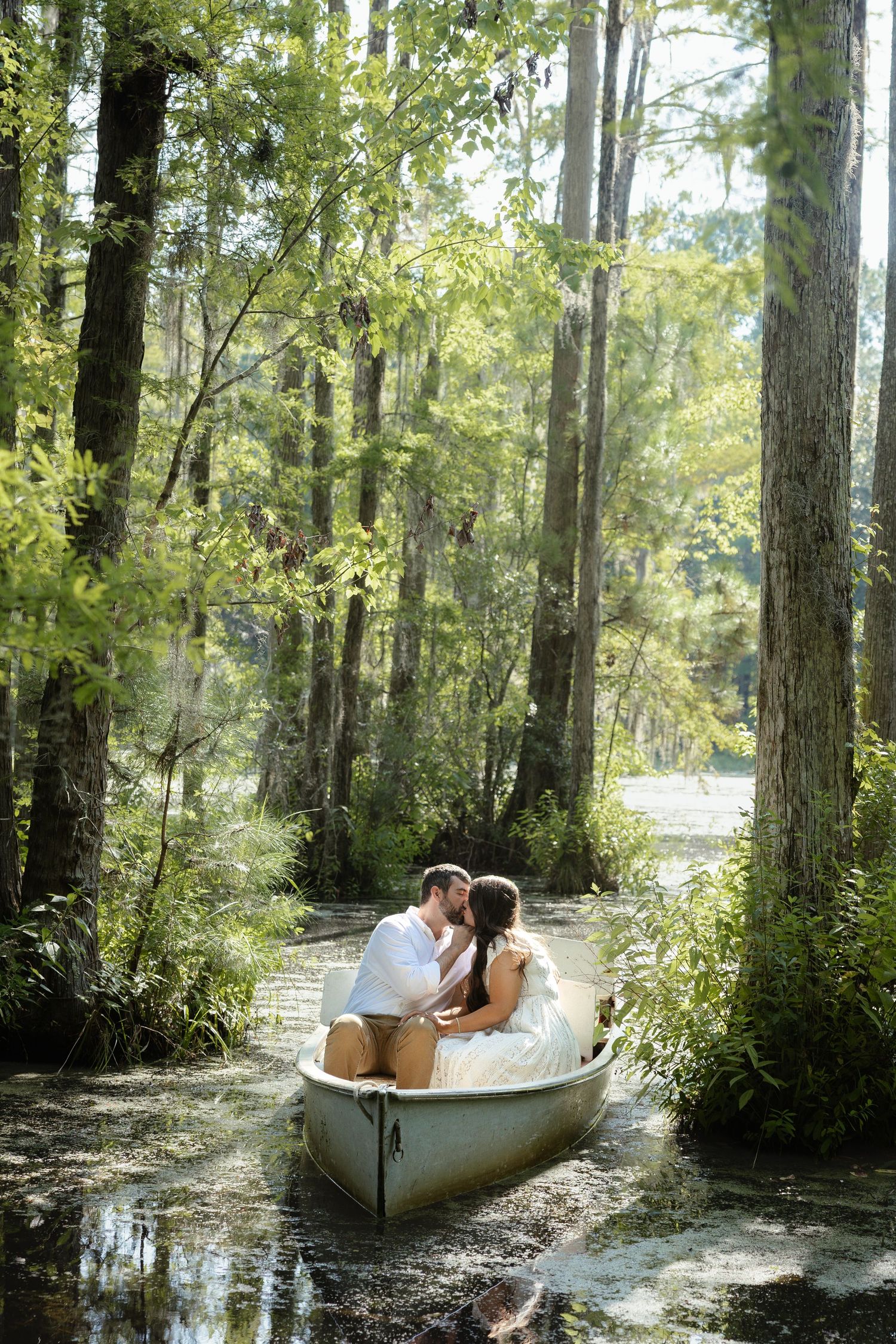 Couple sharing a romantic kiss in a rowboat surrounded by lush green cypress trees in a serene swamp.