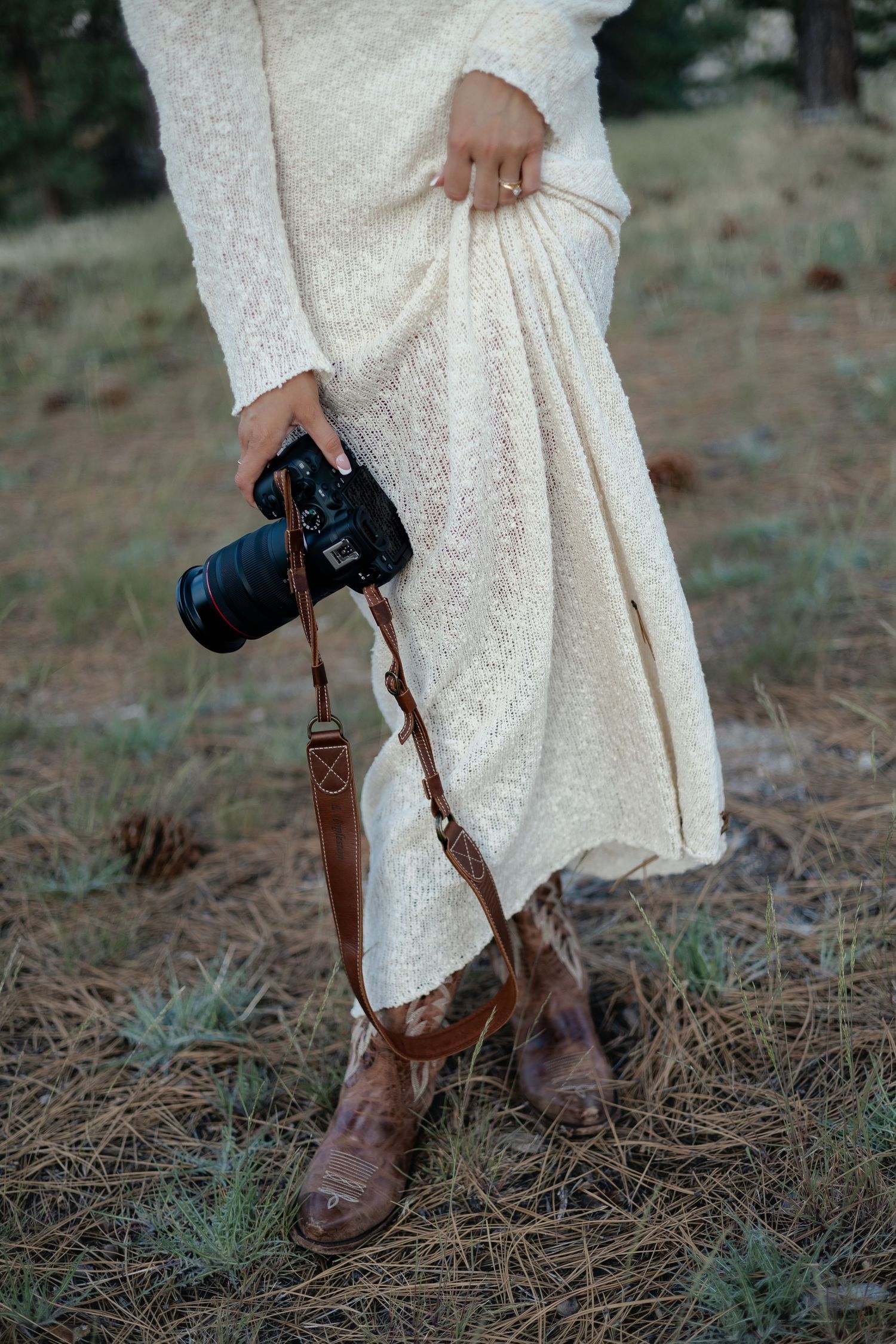 Close-up of a woman in a white lace dress and brown leather boots holding a camera with a leather strap outdoors.