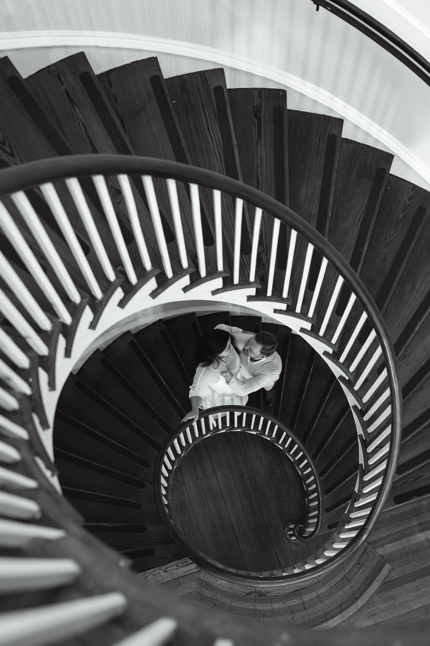 Black and white aerial view of a couple embracing at the center of an elegant spiral staircase.