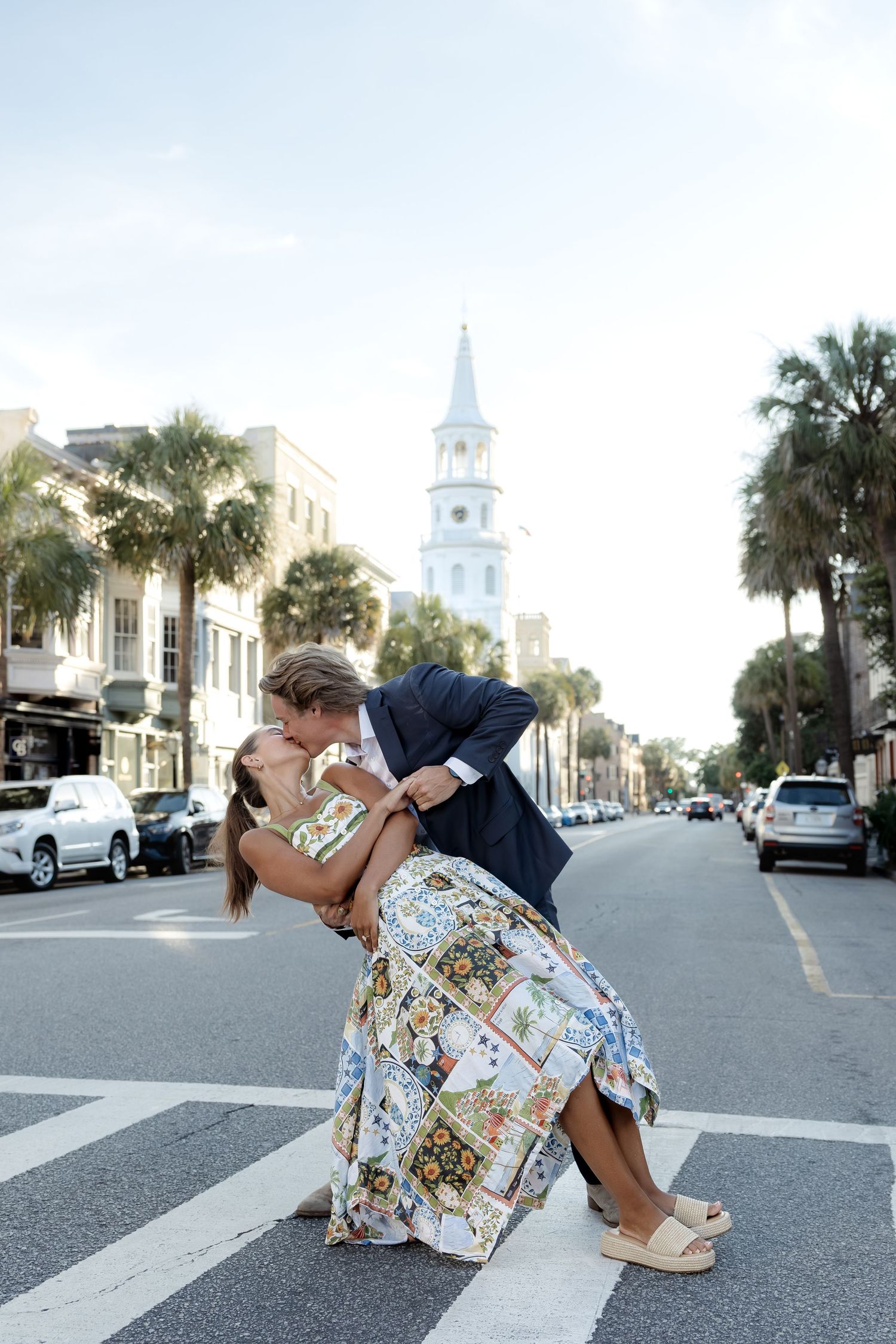 Couple sharing a romantic dip kiss on a street with a white church steeple and palm trees.