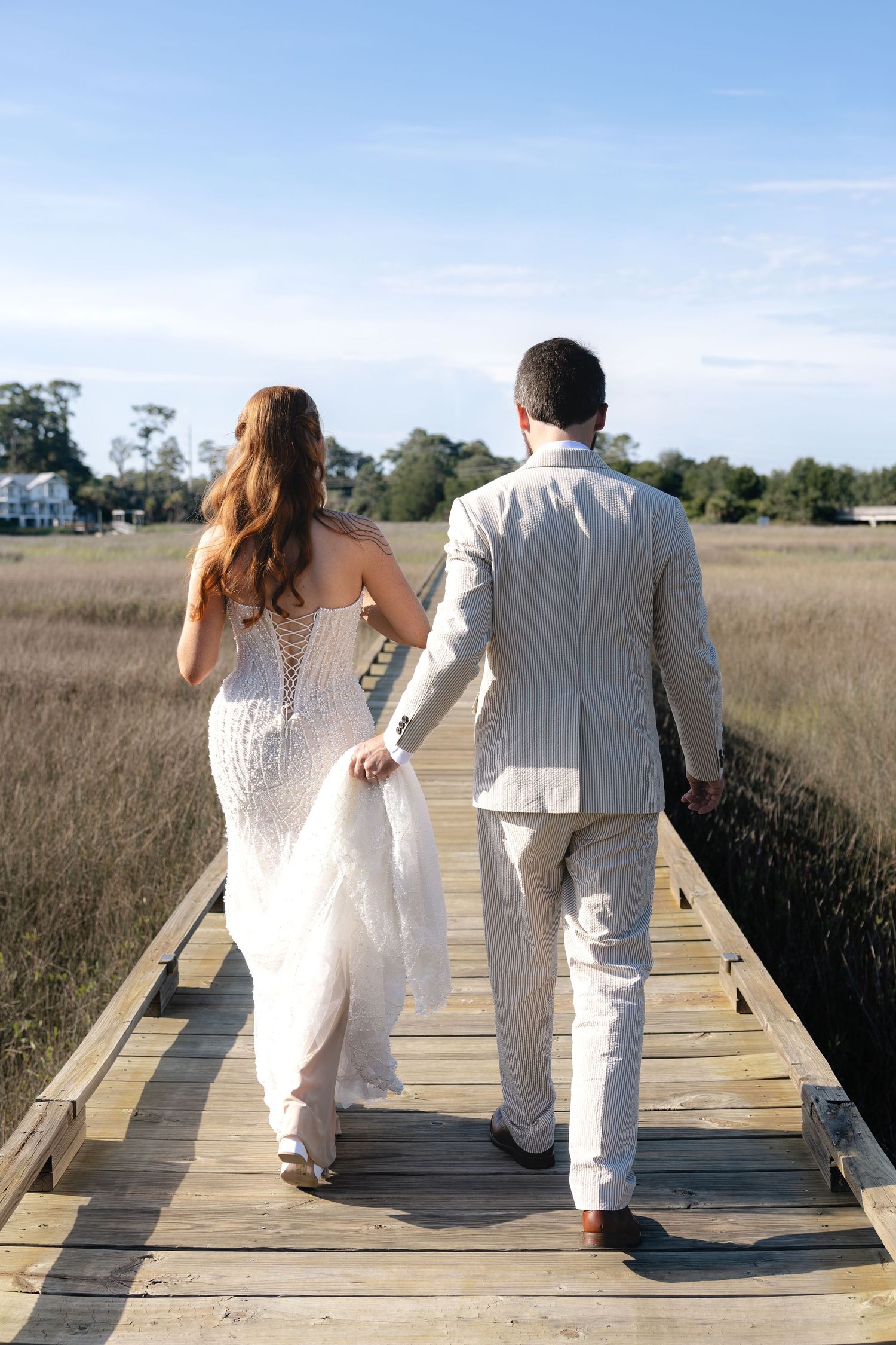 Bride and groom walking hand in hand on a wooden boardwalk through a coastal marsh on their wedding day.