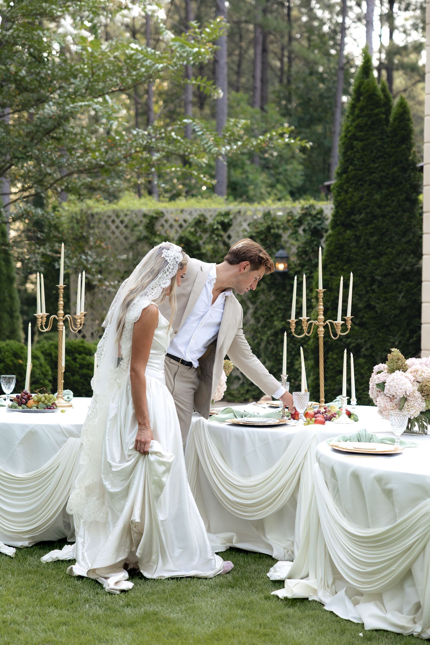 Bride in lace veil and groom arranging an elegant outdoor wedding reception table with candelabras.