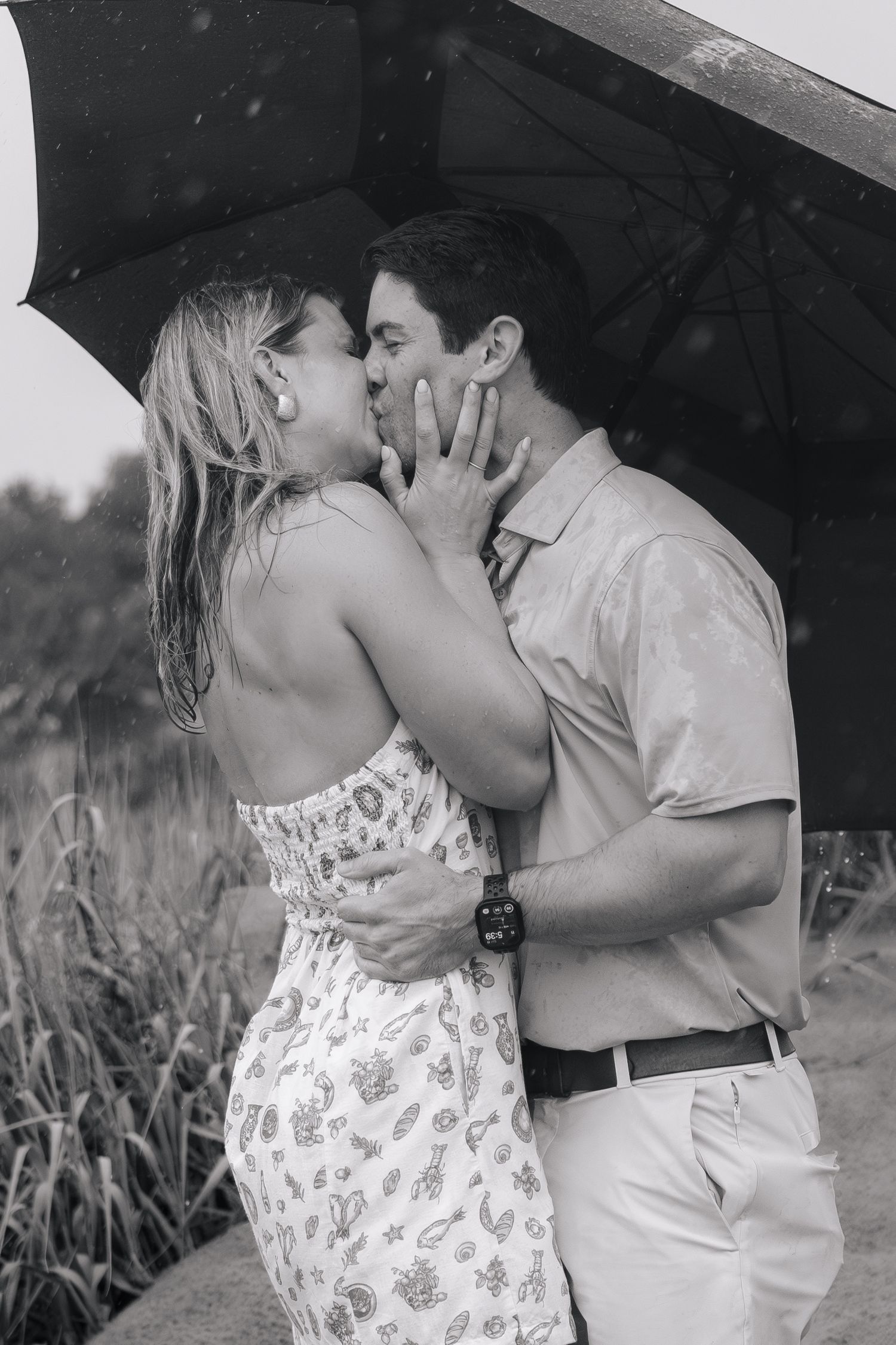 Black and white photo of a couple kissing romantically under an umbrella in the rain outdoors.