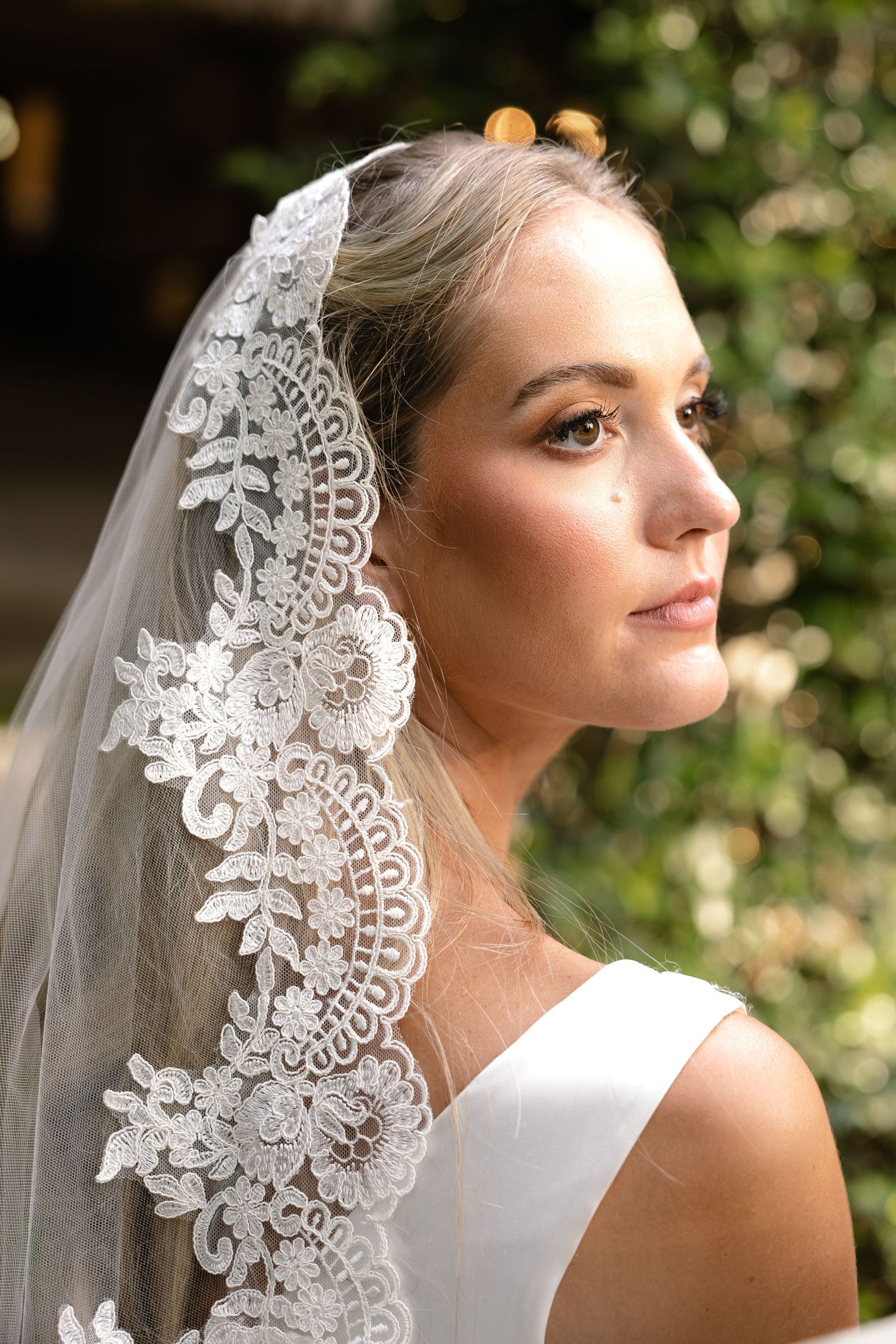 Bride wearing an elegant lace-trimmed wedding veil with floral embroidery, posing outdoors with greenery background.