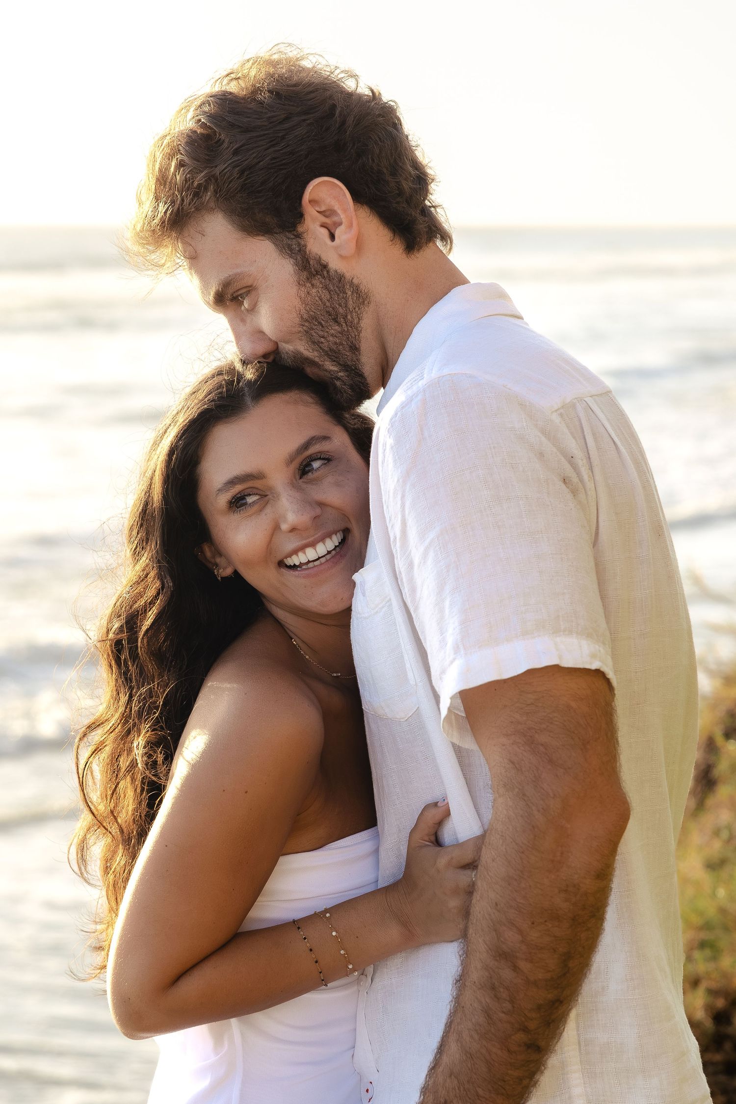 Happy couple embracing on a beach at sunset, both wearing white, woman smiling joyfully as man holds her close.