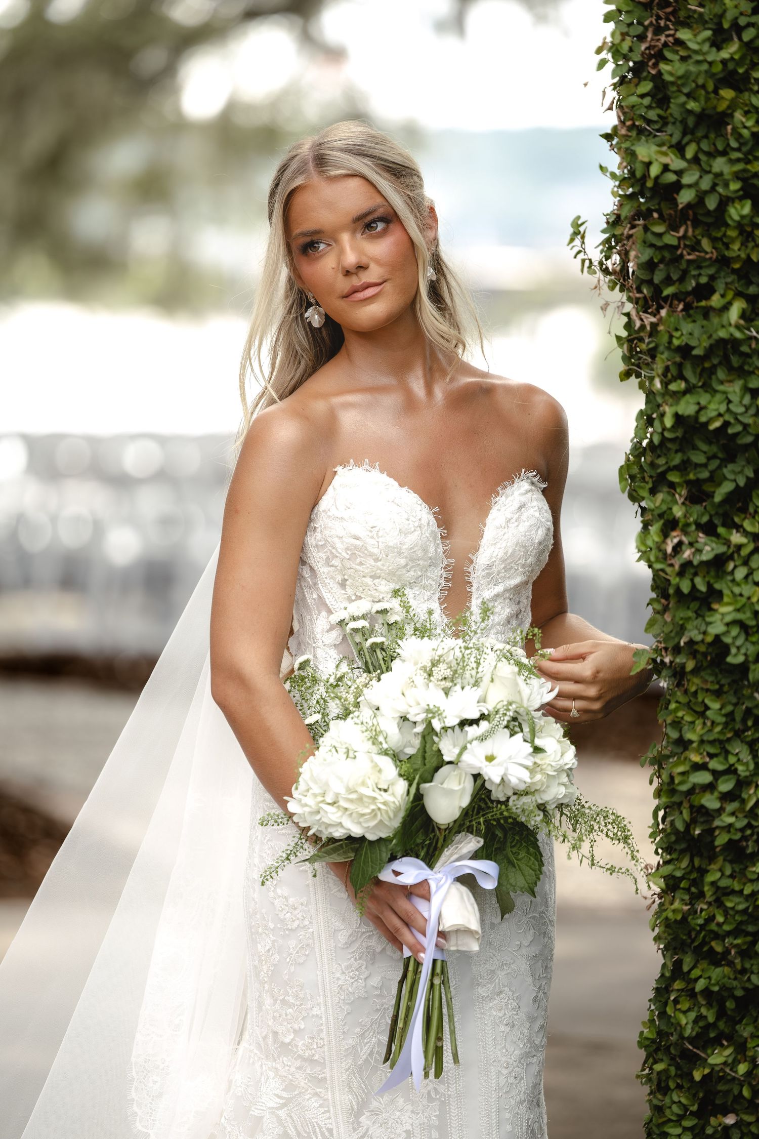 Beautiful bride in a strapless lace wedding gown holding a white floral bouquet with a veil outdoors.