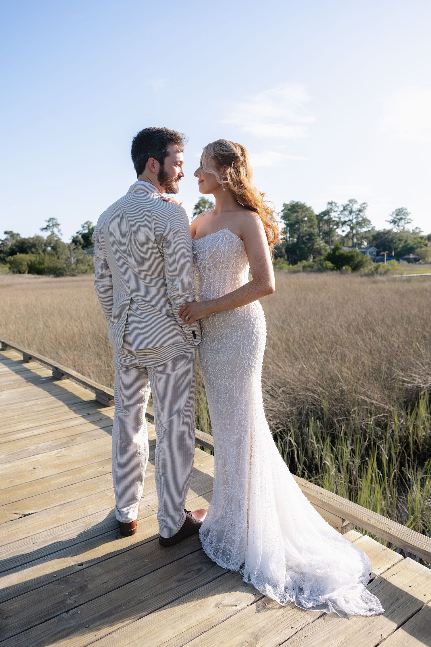 Bride and groom in wedding attire standing on wooden boardwalk overlooking golden marsh grasses under blue sky.