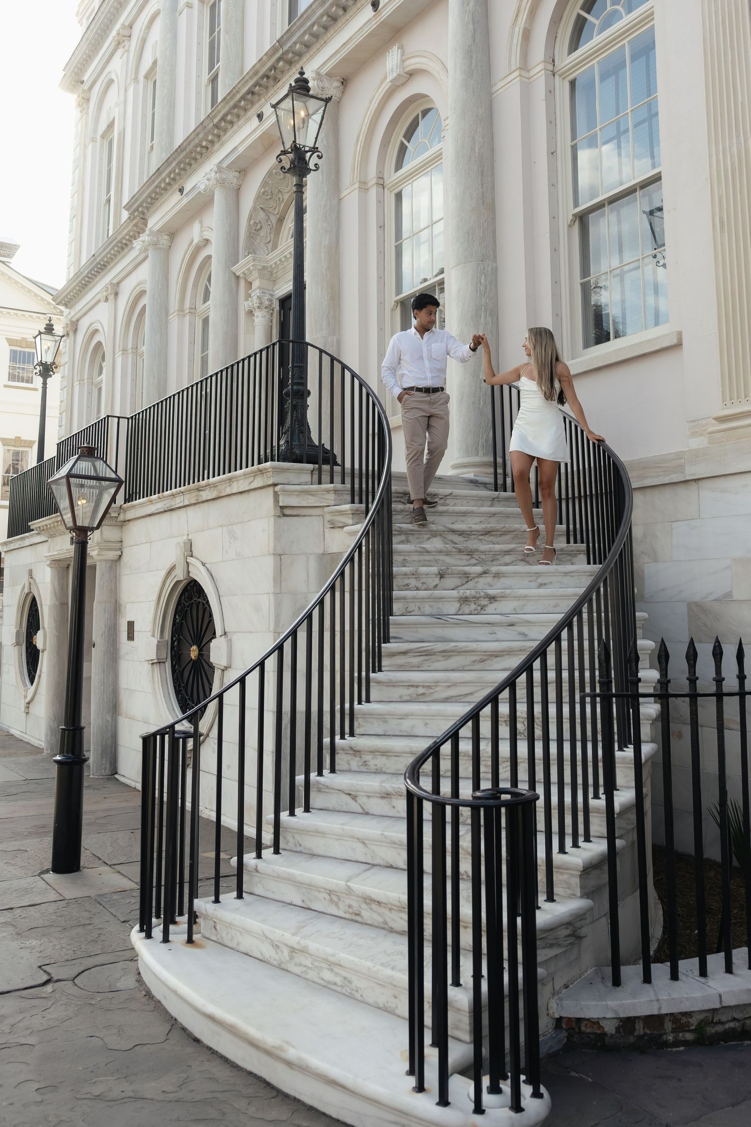Couple holding hands while descending elegant curved marble stairs of a grand white historic building.