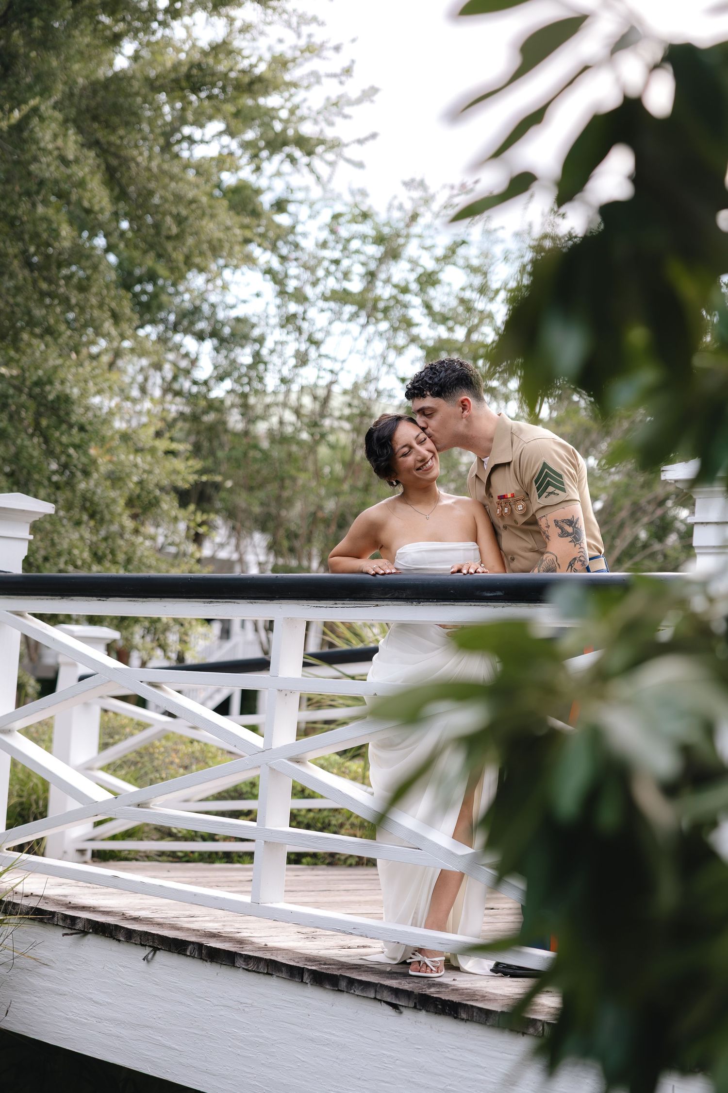 Couple sharing a romantic kiss on a white wooden bridge surrounded by lush green trees in an outdoor garden setting.