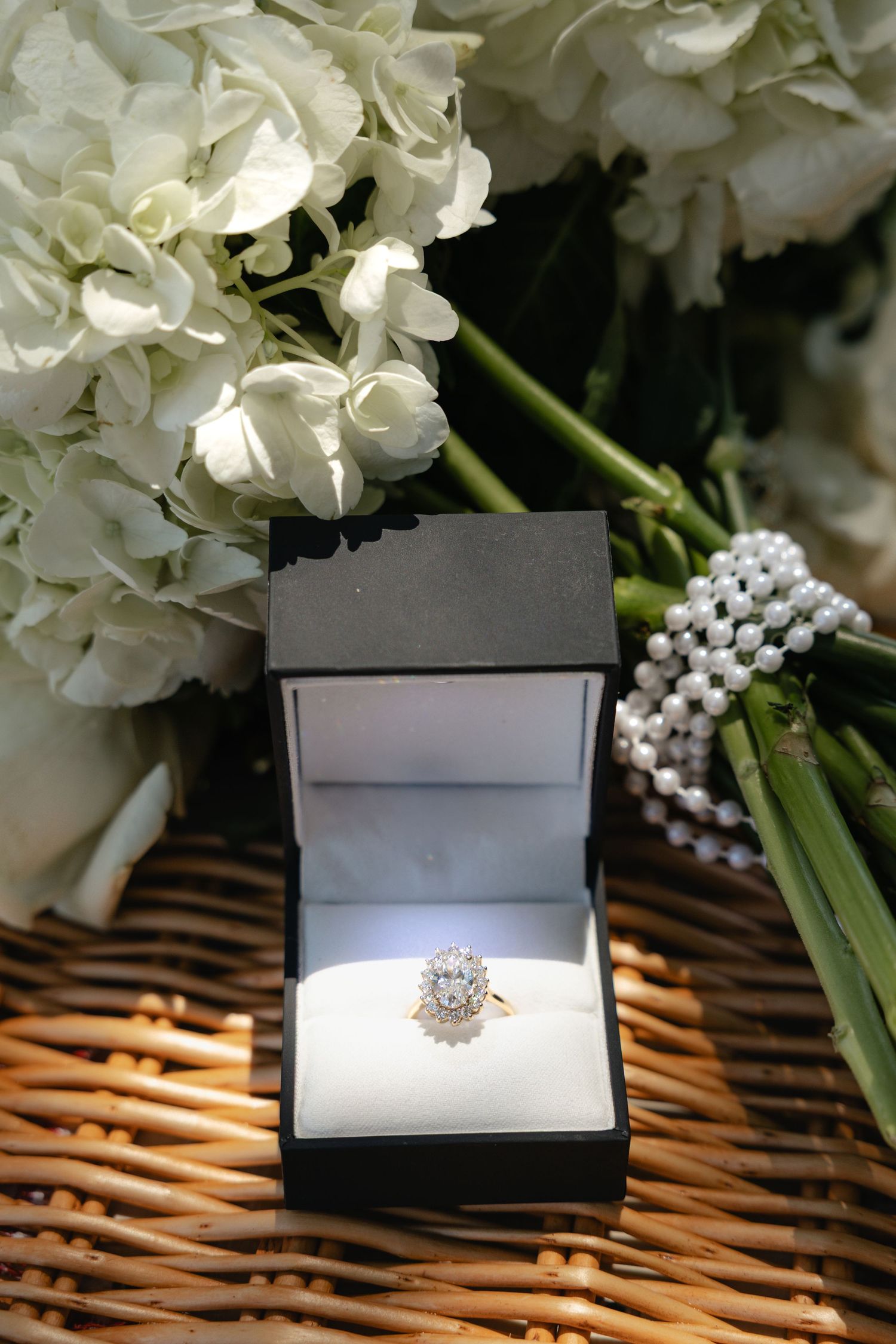 Diamond halo engagement ring in black jewelry box surrounded by white hydrangeas and pearl bracelet.