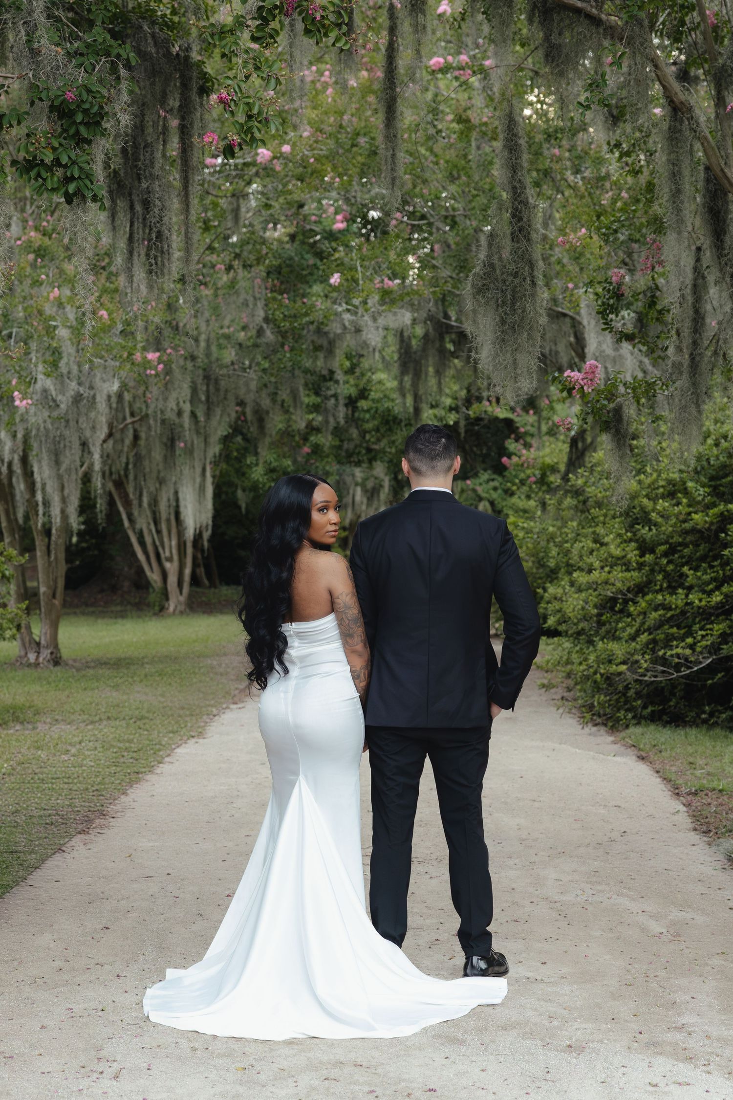 Bride in white strapless gown and groom in black suit stand on moss-draped path at outdoor wedding.