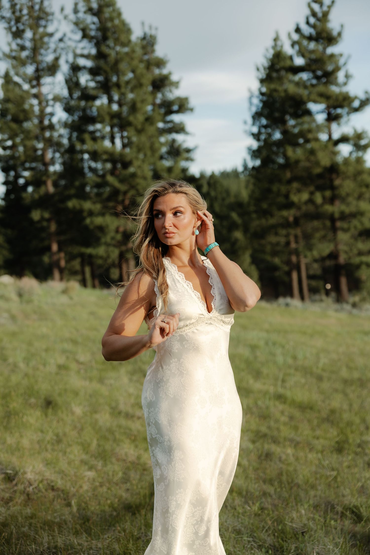 Blonde woman in elegant white lace gown posing in a meadow with tall pine trees in the background at sunset.
