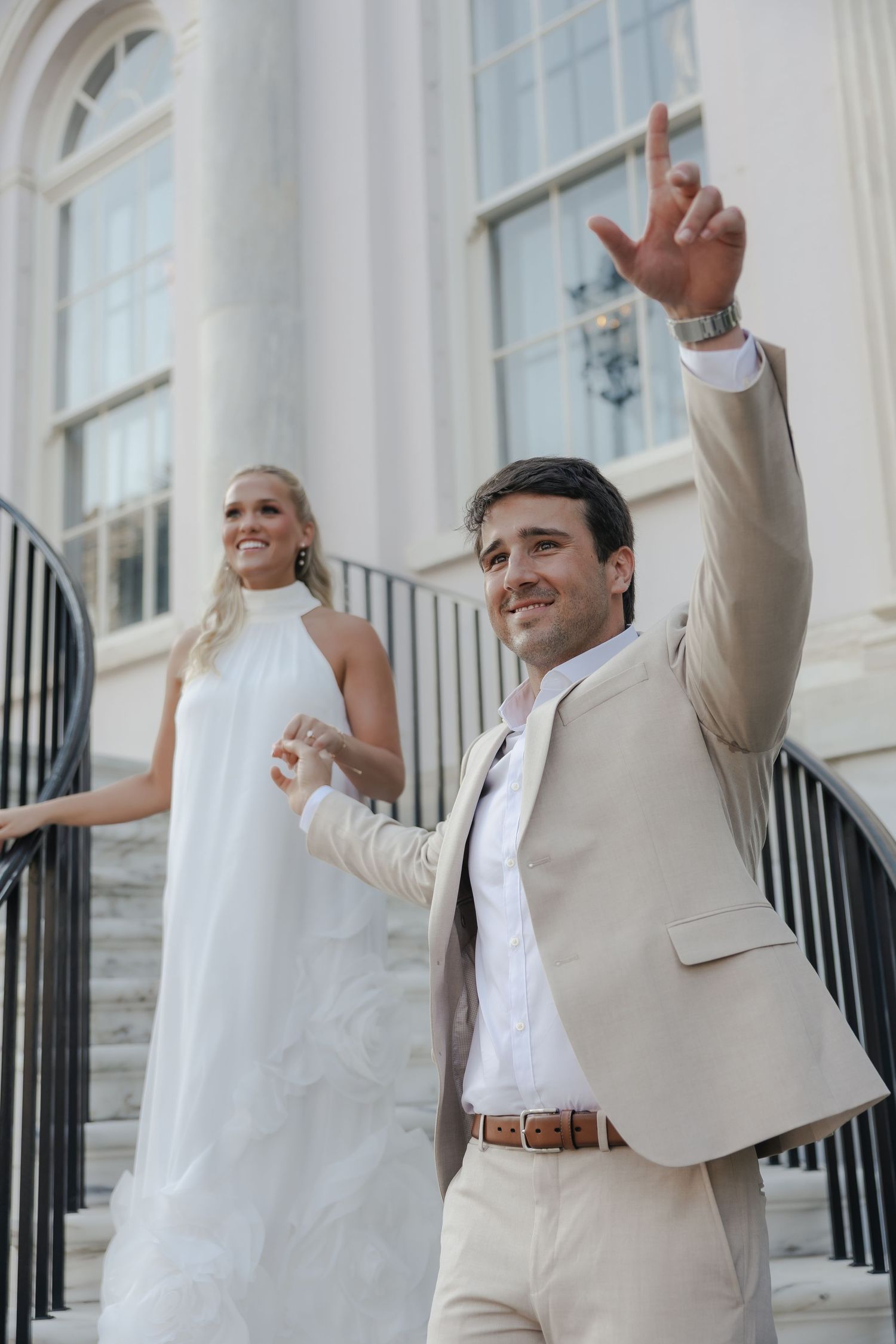 Smiling groom in tan suit raises hand while bride in white halter gown stands on elegant building steps.
