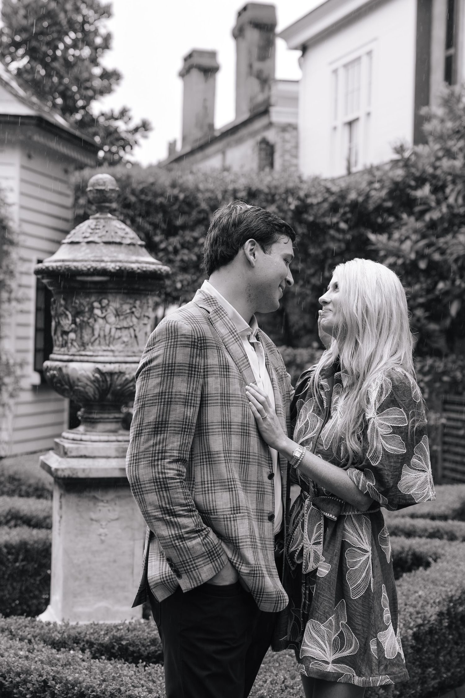 Black and white photo of a couple sharing a romantic moment in an elegant garden with hedges and a historic mansion backdrop.