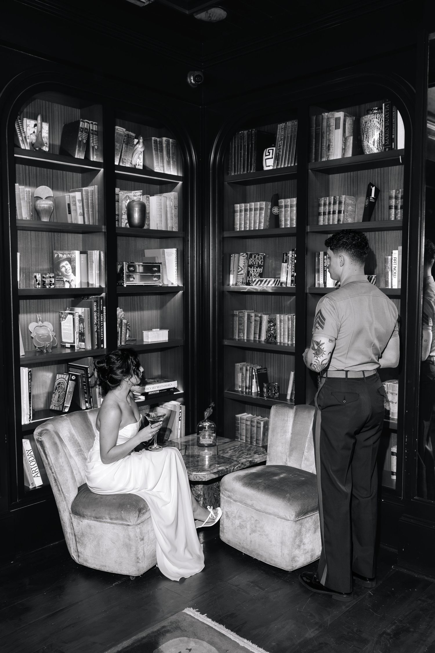 Black and white photo of a bride sitting in a velvet chair reading in an elegant library while groom stands nearby.