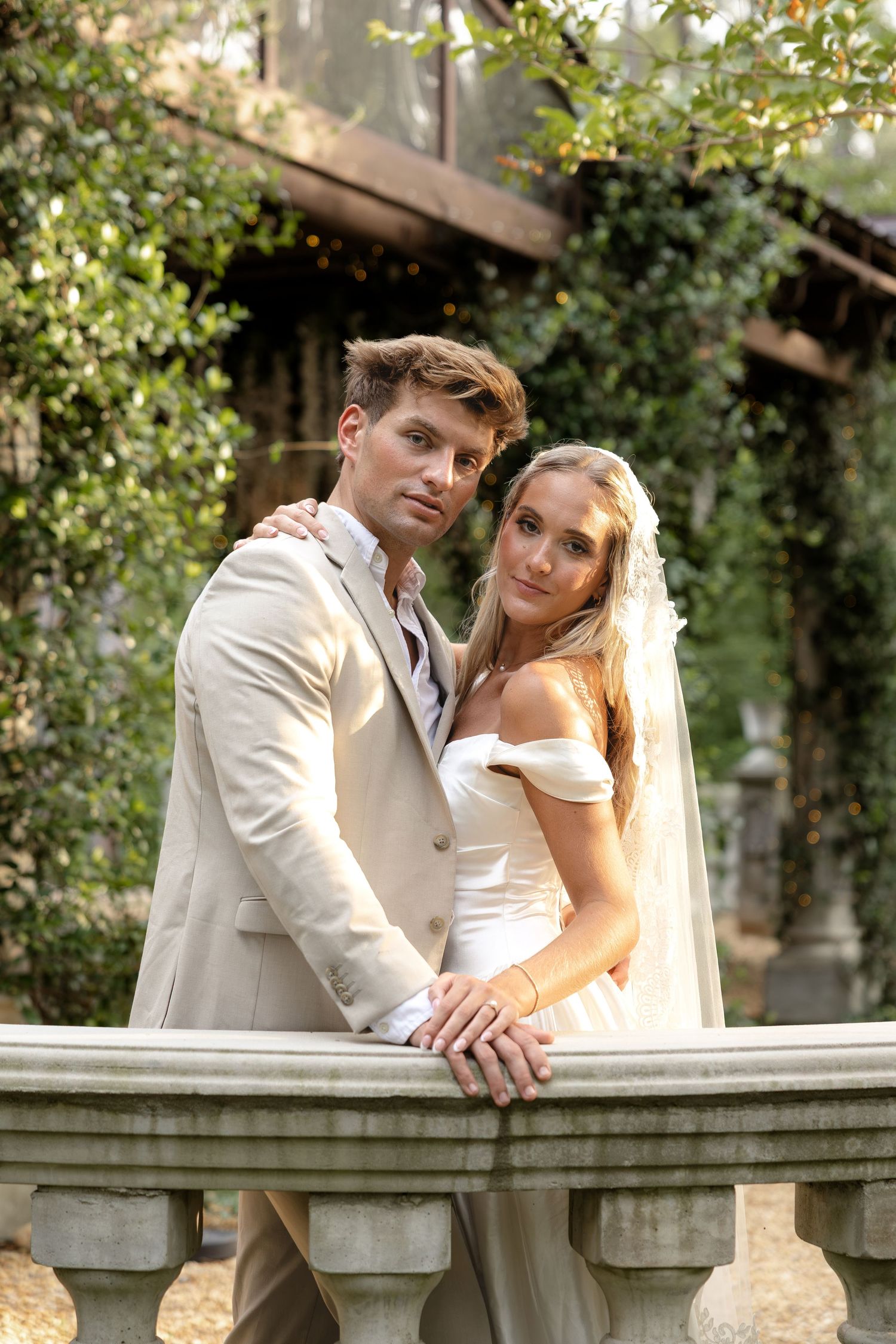Elegant bride and groom posing together at a stone balustrade surrounded by lush greenery on their wedding day.