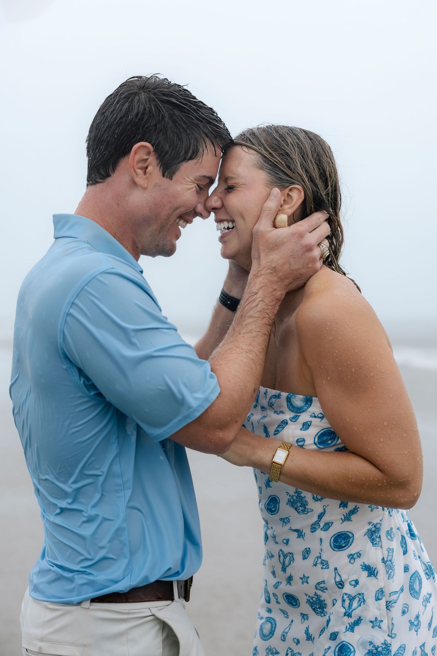 Joyful couple with wet hair laughing and embracing on a misty beach during an engagement session.