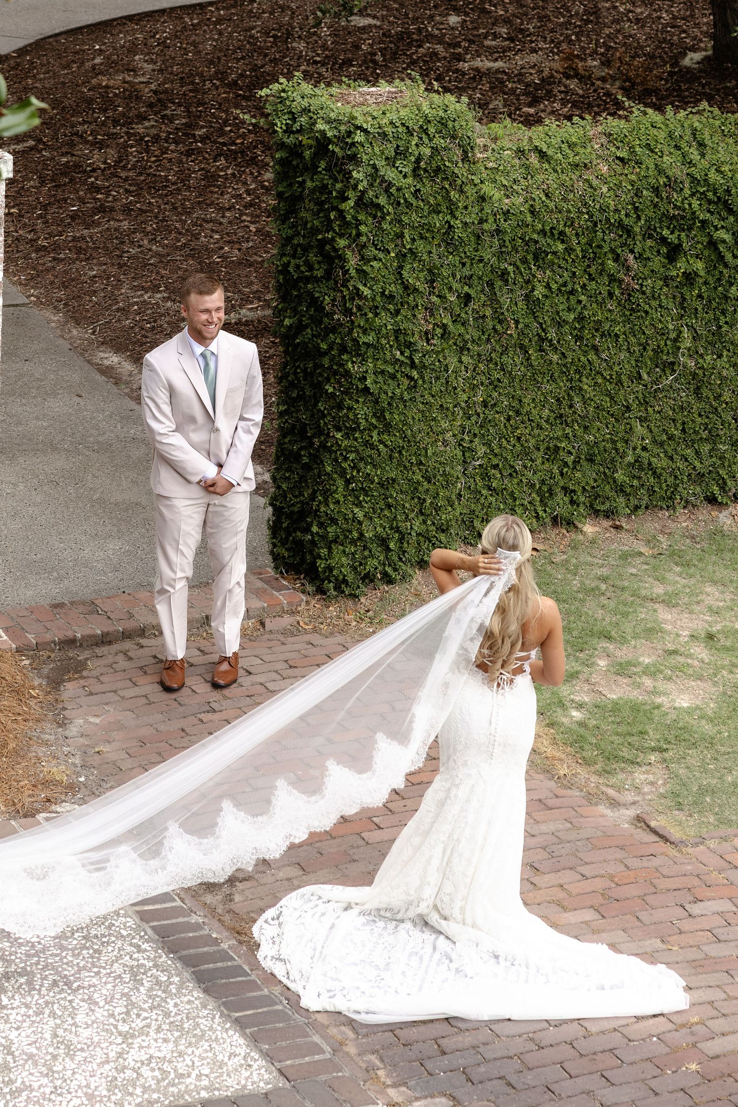 Bride in lace gown with long veil faces groom in tan suit during outdoor first look on brick path.