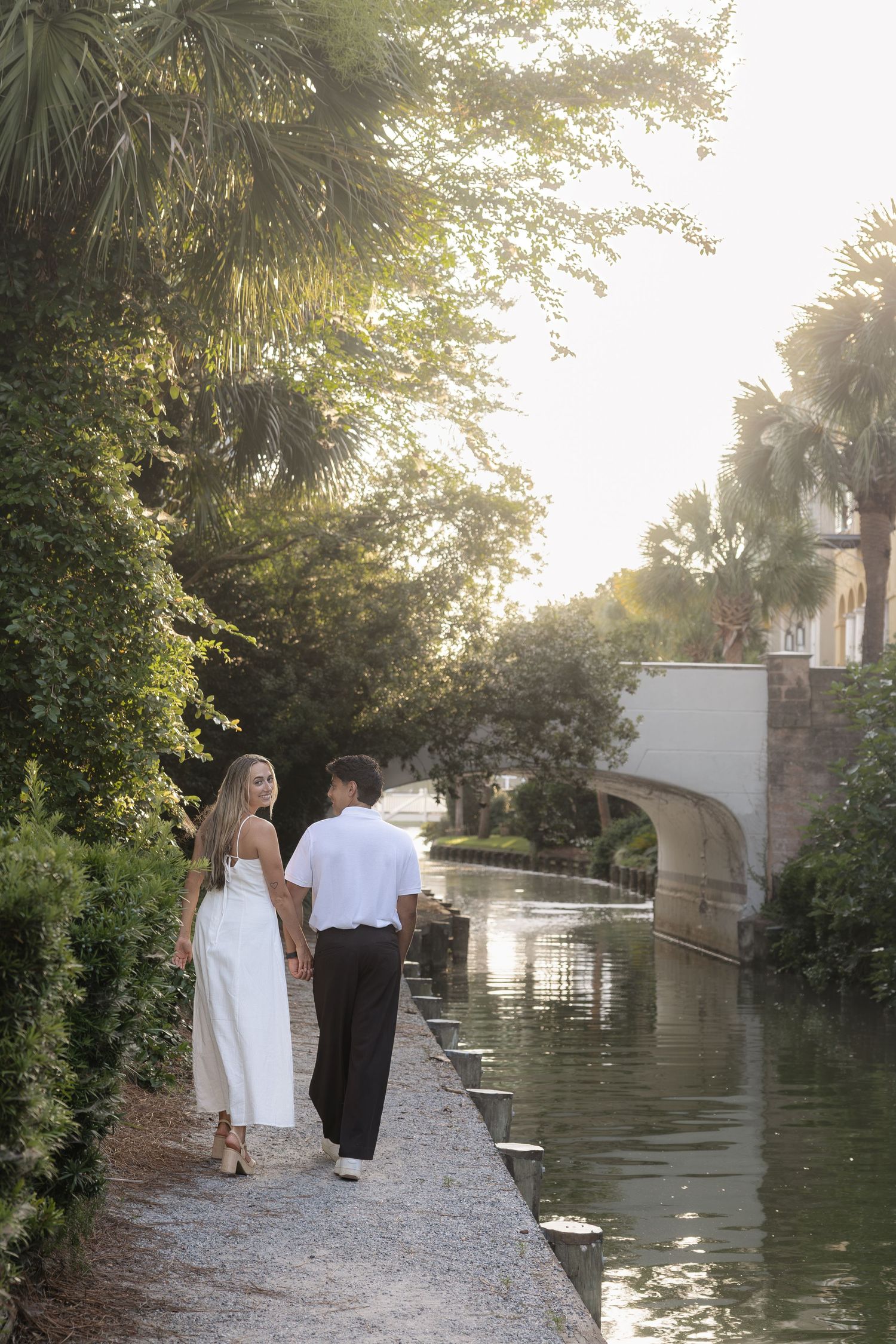 Couple walking hand in hand along a scenic waterway path surrounded by lush palm trees in golden sunlight.