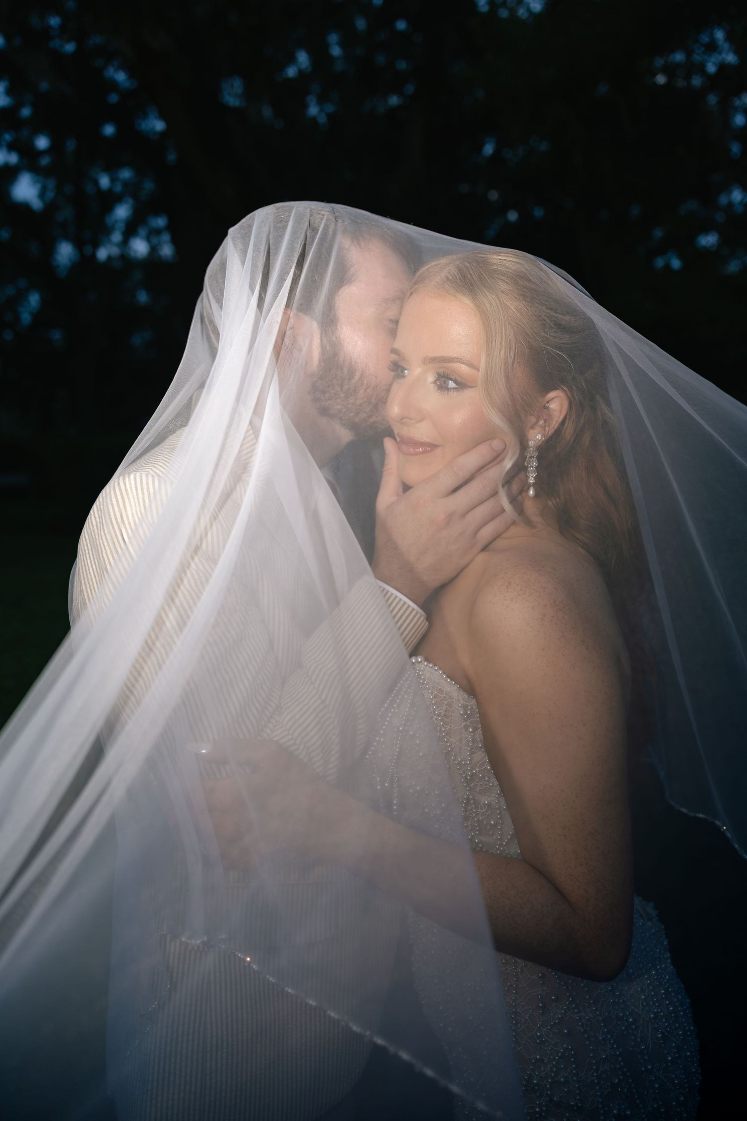 Bride and groom share romantic kiss under wedding veil at night, bride wearing strapless gown and chandelier earrings.