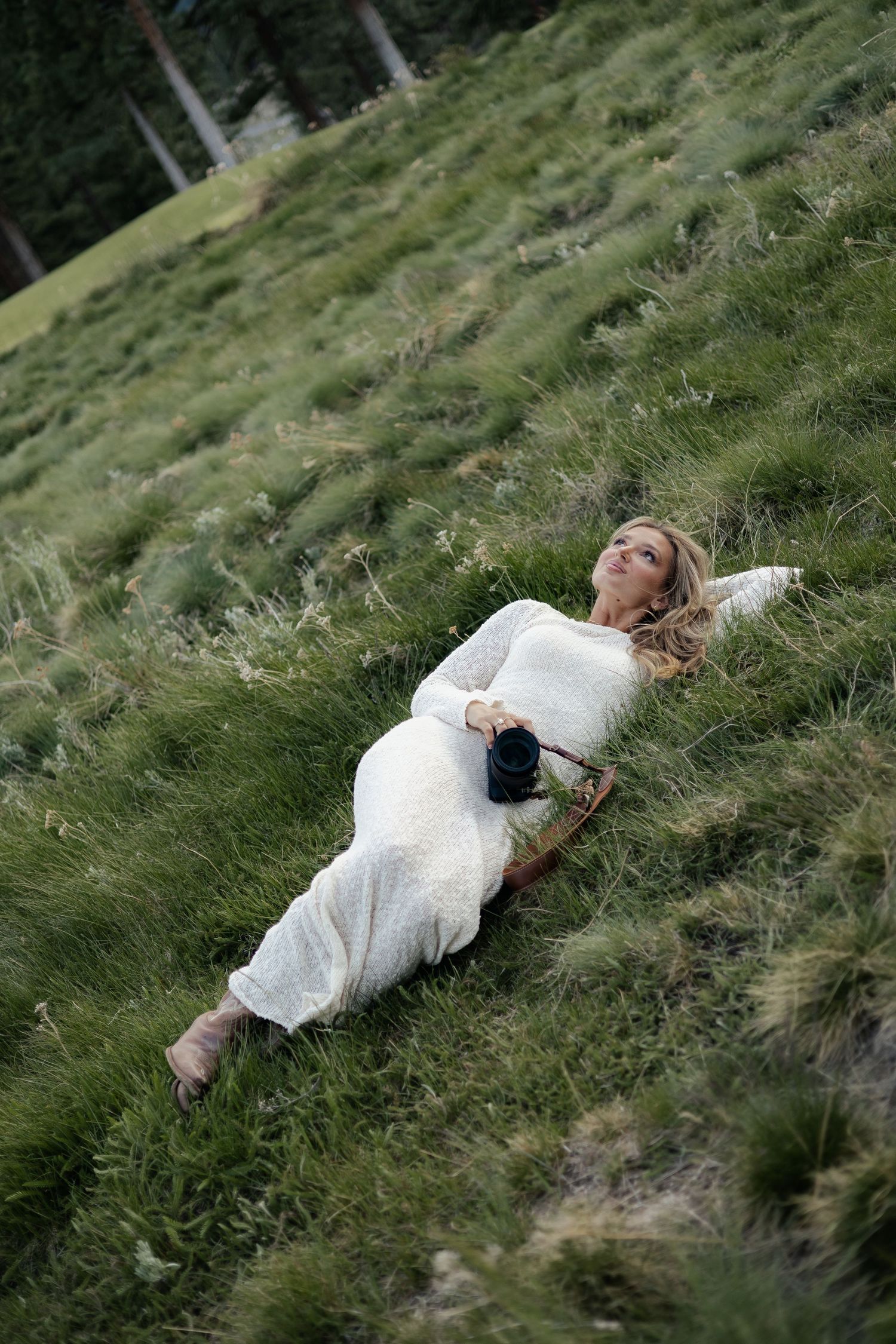 Woman in white dress lying on a grassy hillside holding a camera, gazing up at the sky near pine trees.