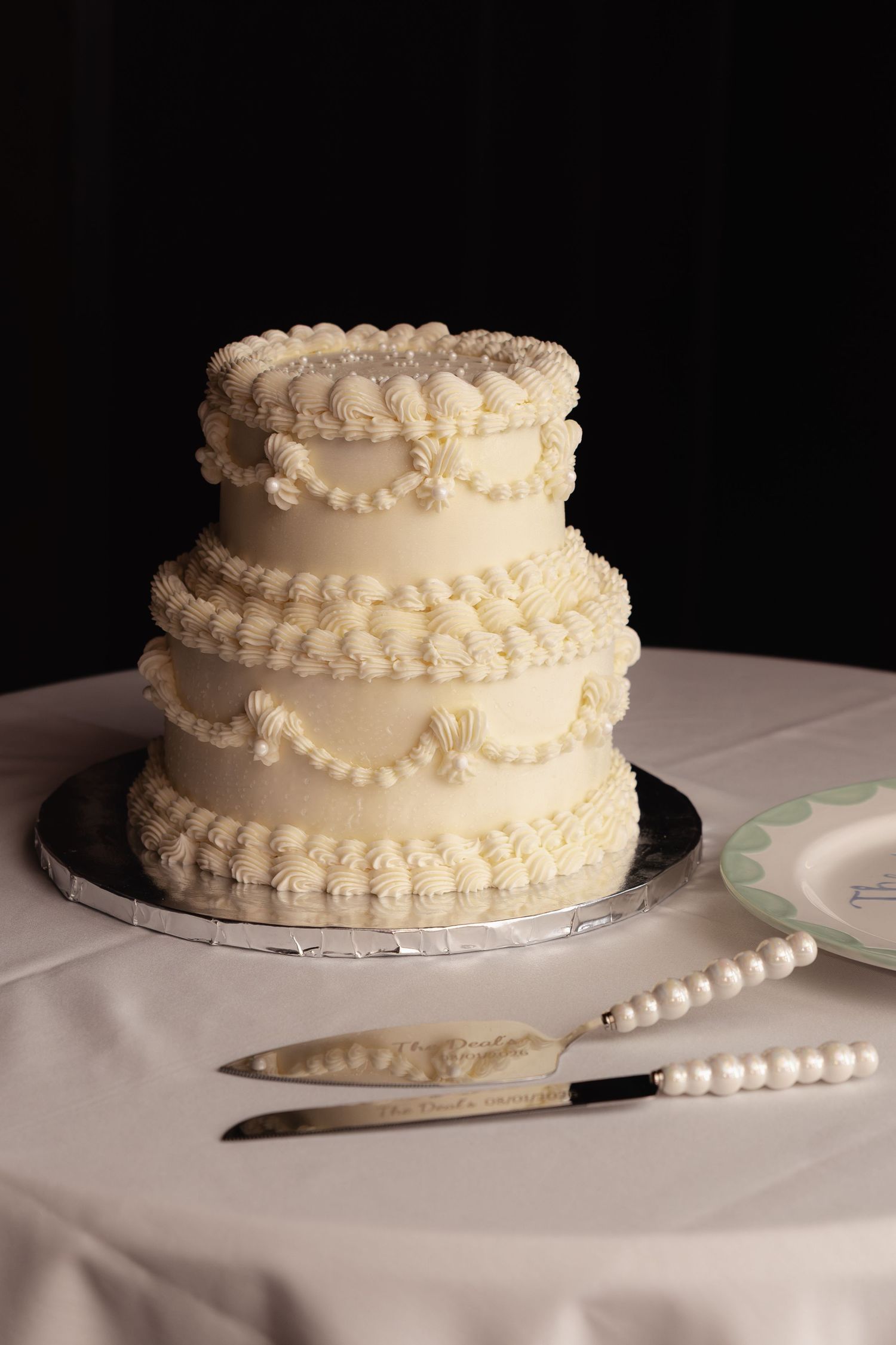 Elegant three-tier white wedding cake with pearl piping decorations on silver stand with cake cutting knife set.