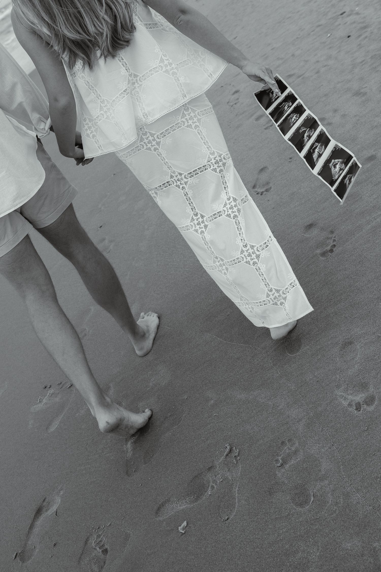 Black and white aerial view of a couple walking on sand holding ultrasound photos.