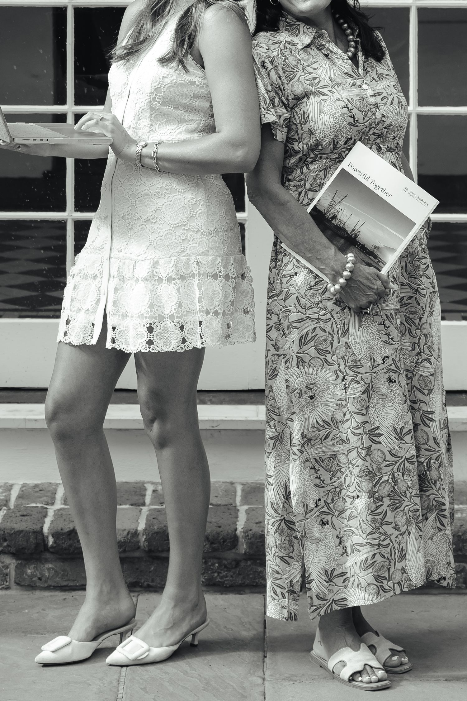 Black and white photo of two women in summer dresses, one holding a book, standing outside a building.