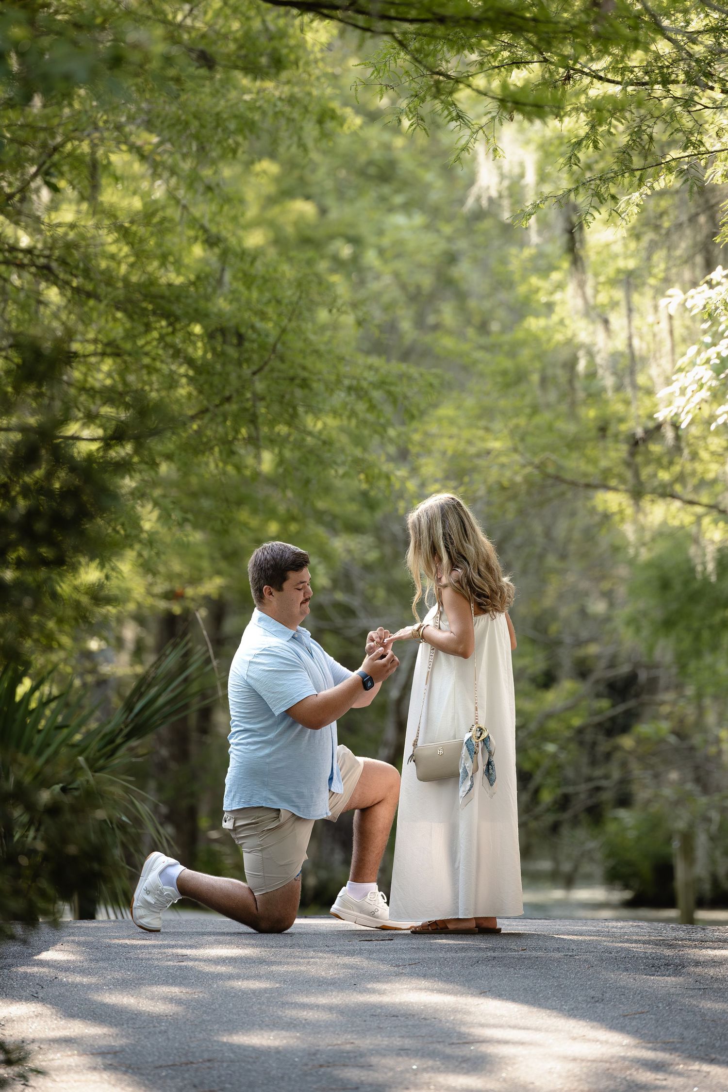 Man kneeling on one knee proposing to woman in white dress on a tree-lined path in a lush green park.