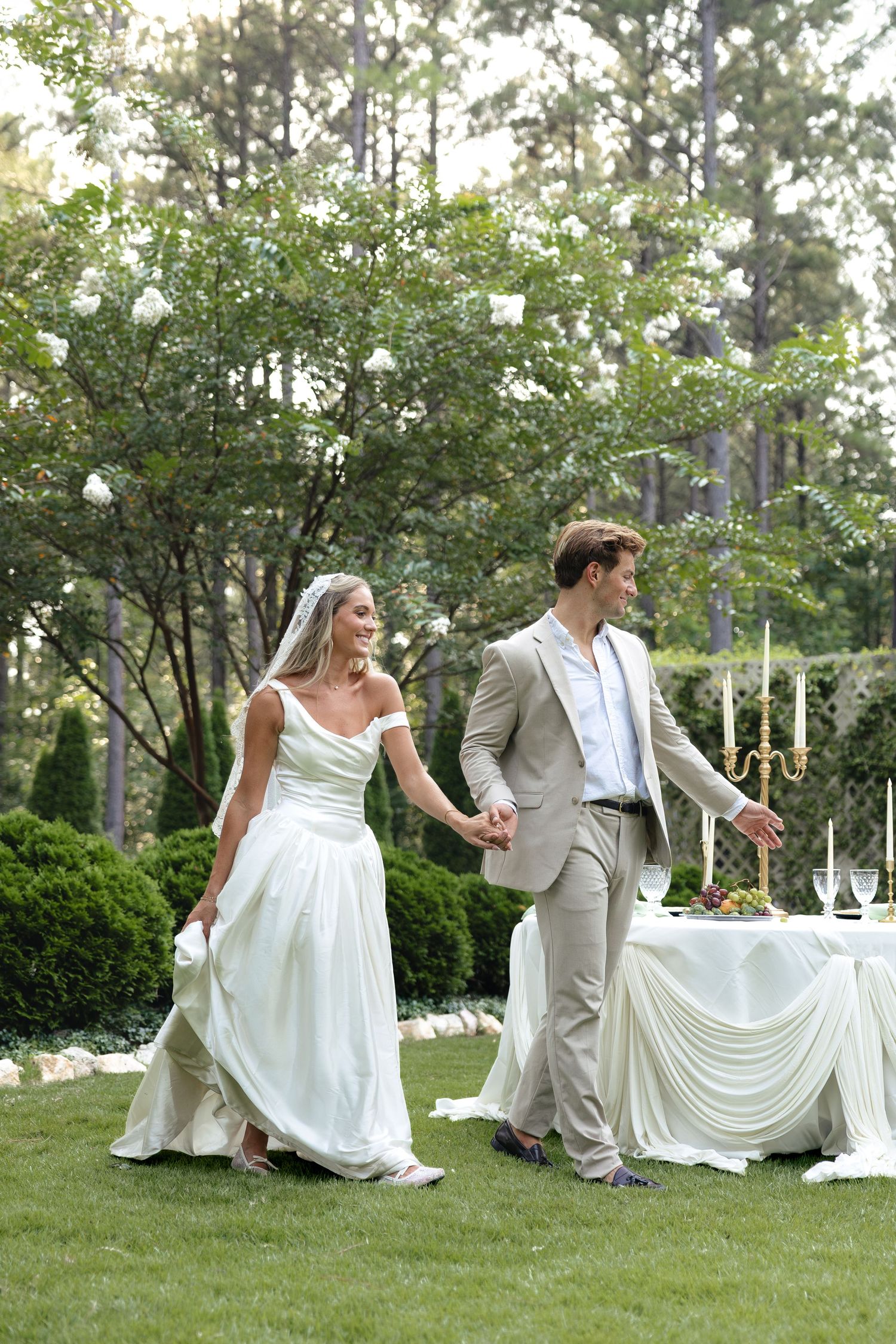 A bride and groom walk hand in hand on a green lawn during an outdoor garden wedding ceremony.