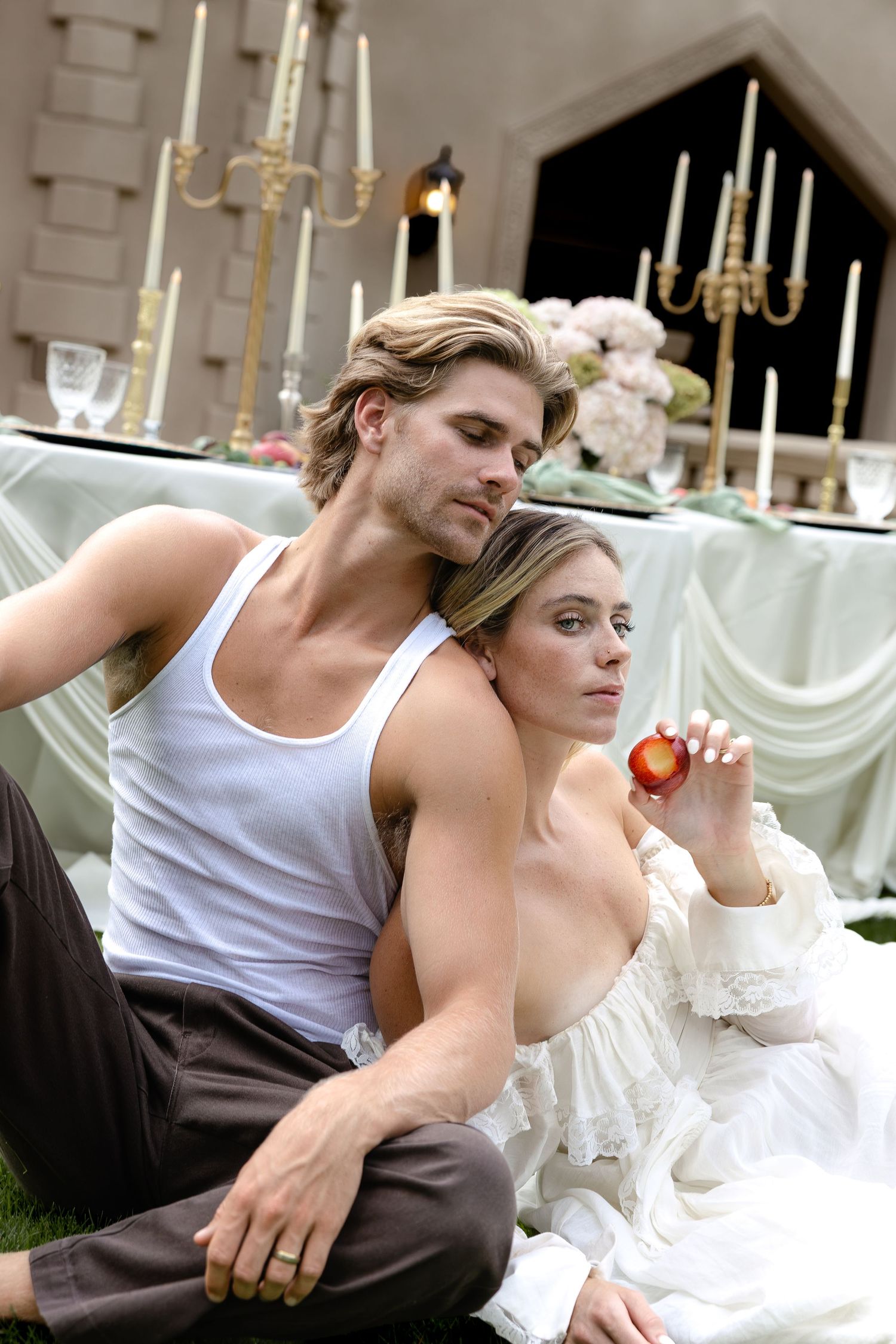 A couple sits back-to-back outdoors near a candlelit table, the woman in white holding a peach.