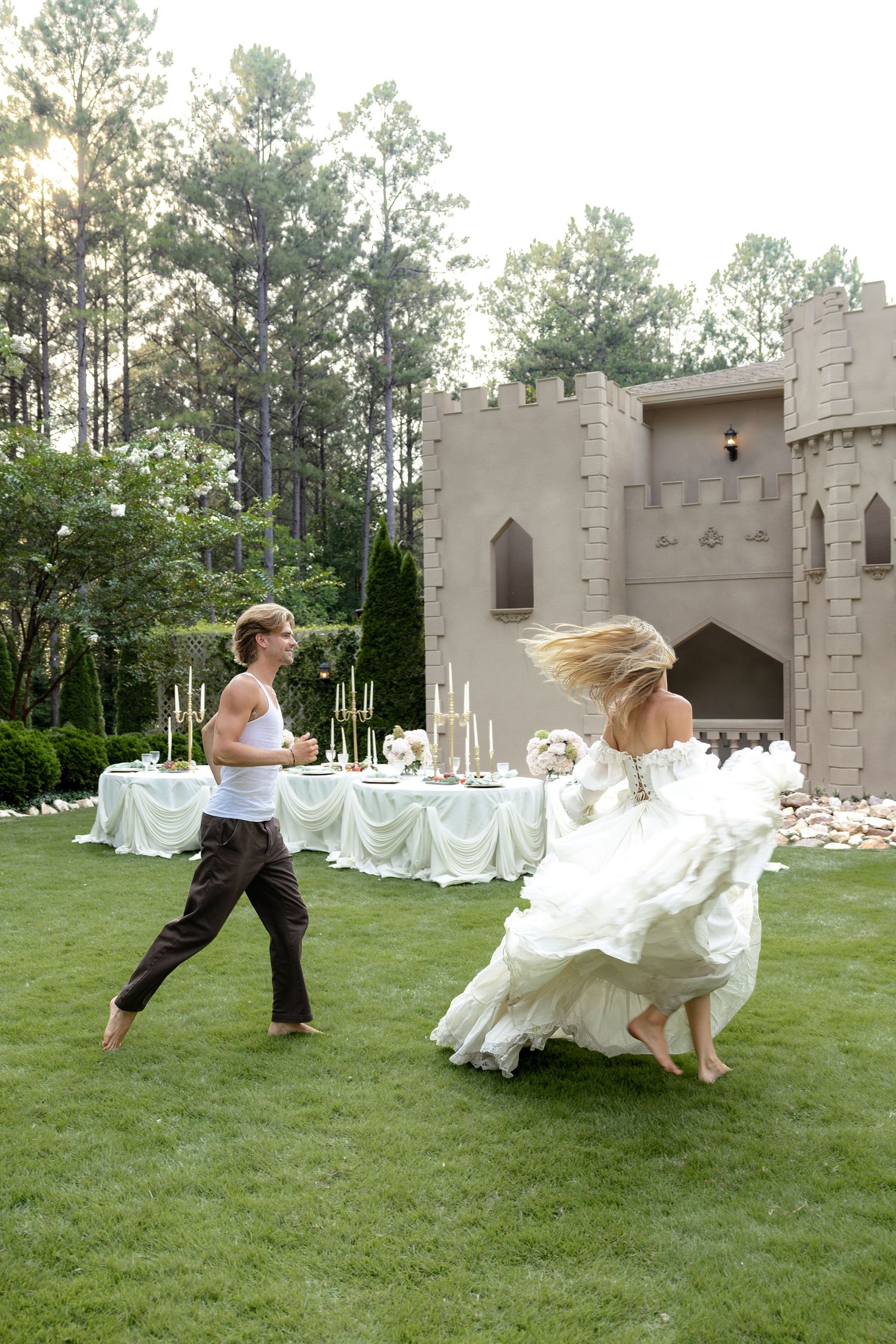Bride in white gown and groom in casual attire dance barefoot on green lawn beside castle backdrop.