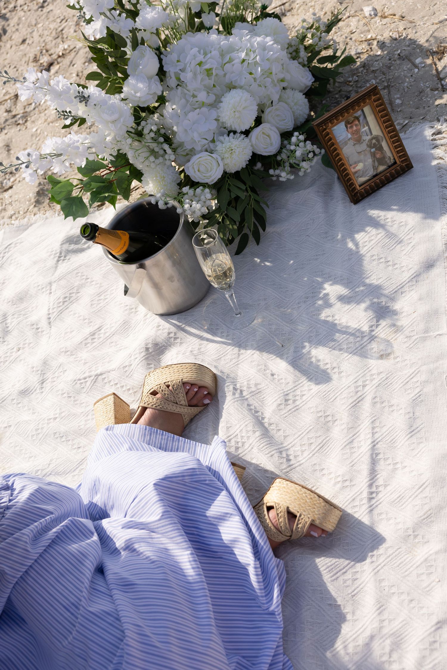 Beach picnic setup with champagne, white flowers, framed photo on white blanket.