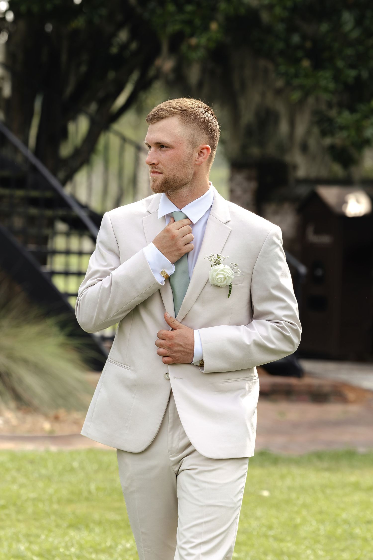 Groom in beige suit with sage green tie and white boutonniere adjusts his jacket outdoors at a wedding.