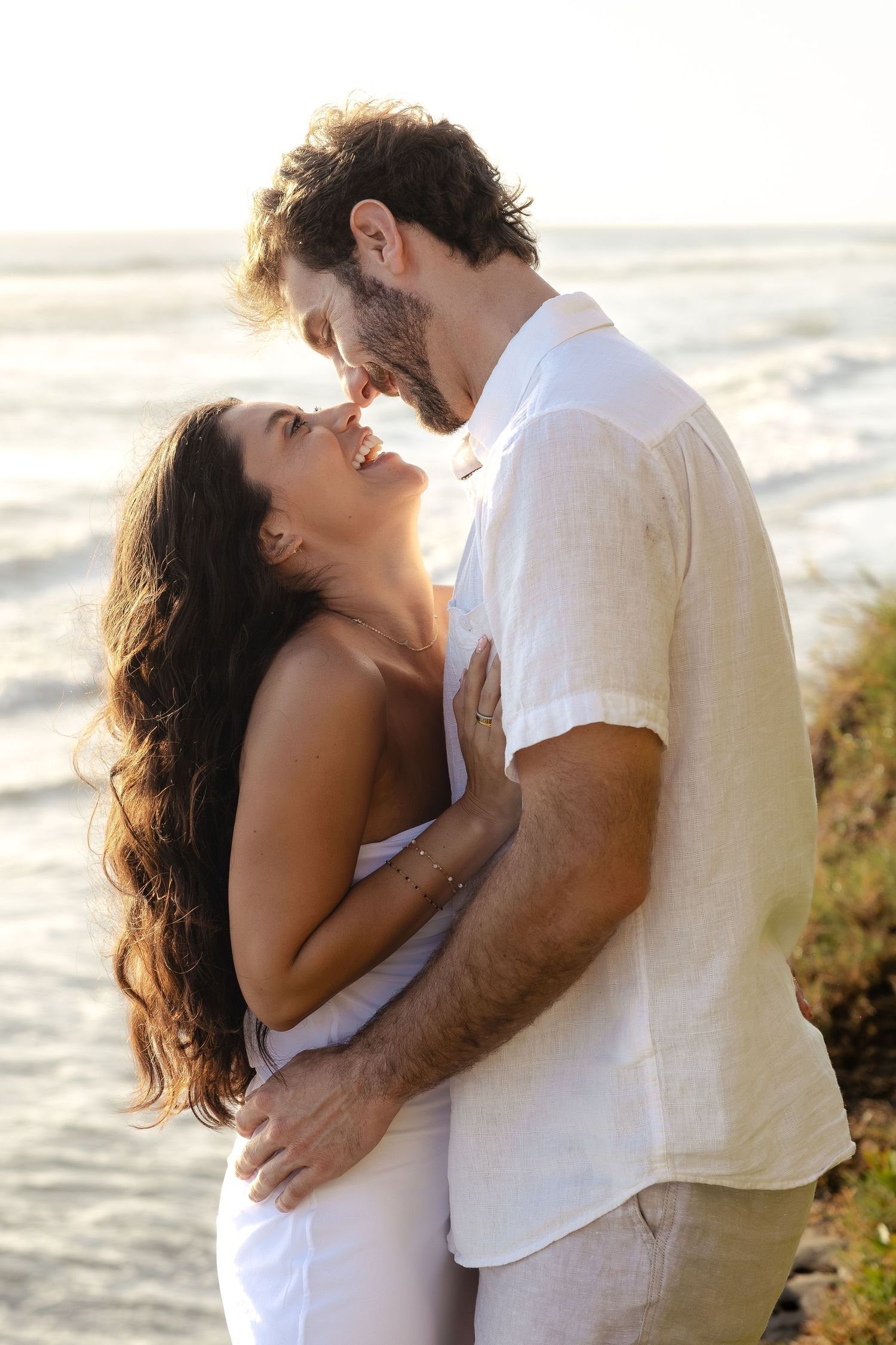 Romantic couple embracing and smiling at each other on a beach at sunset, wearing white outfits.