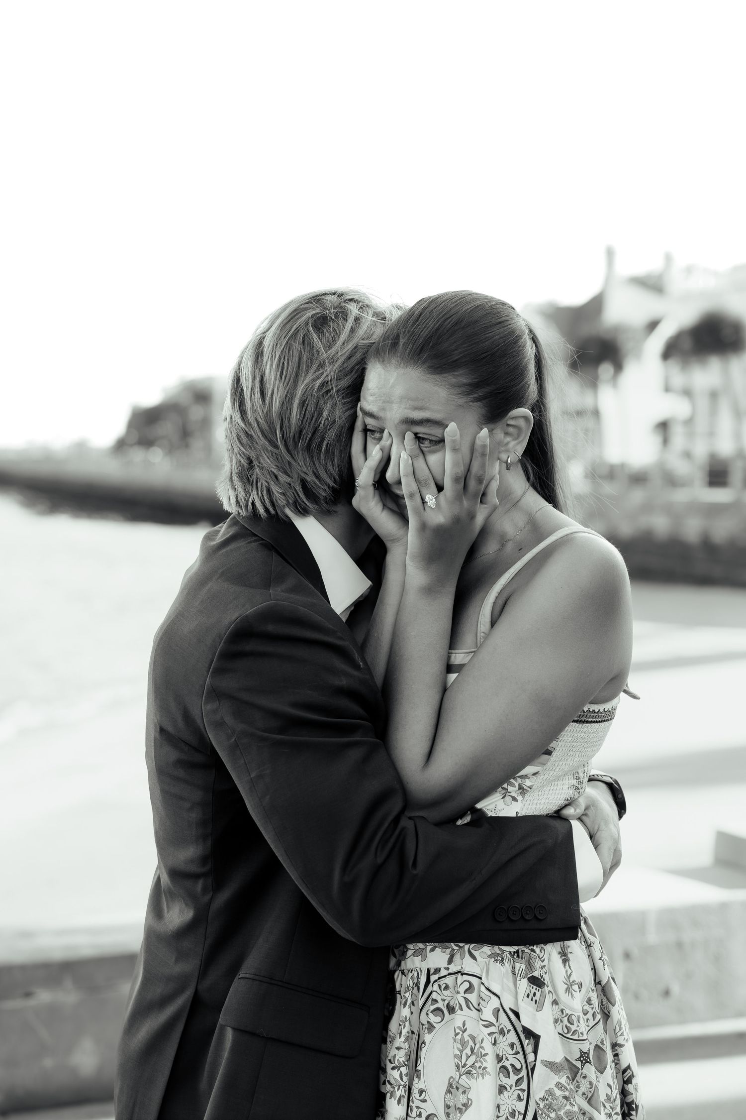Emotional black and white photo of woman covering her face with hands after marriage proposal by waterfront.