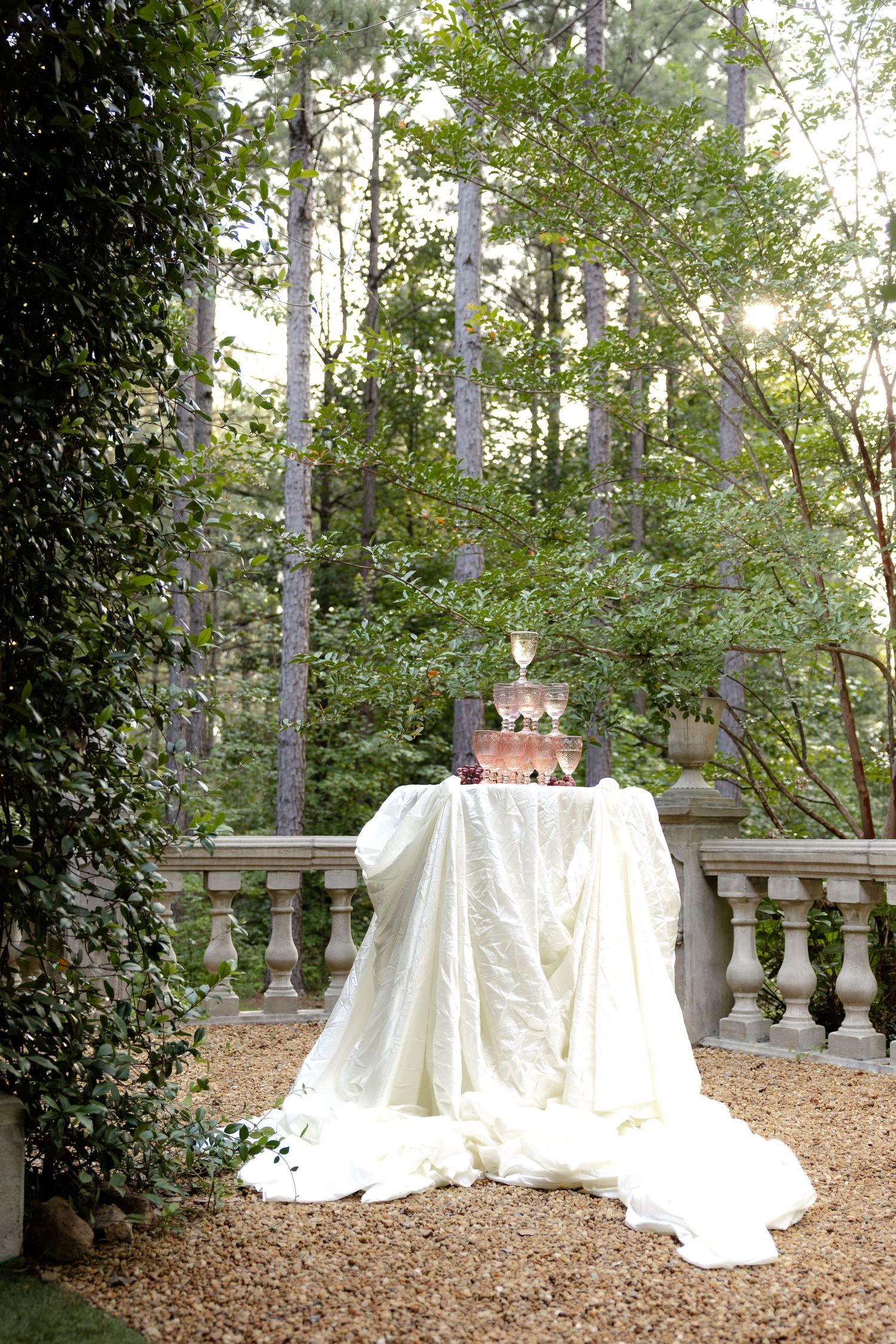 Outdoor wedding champagne tower on draped white linen table surrounded by lush green trees.