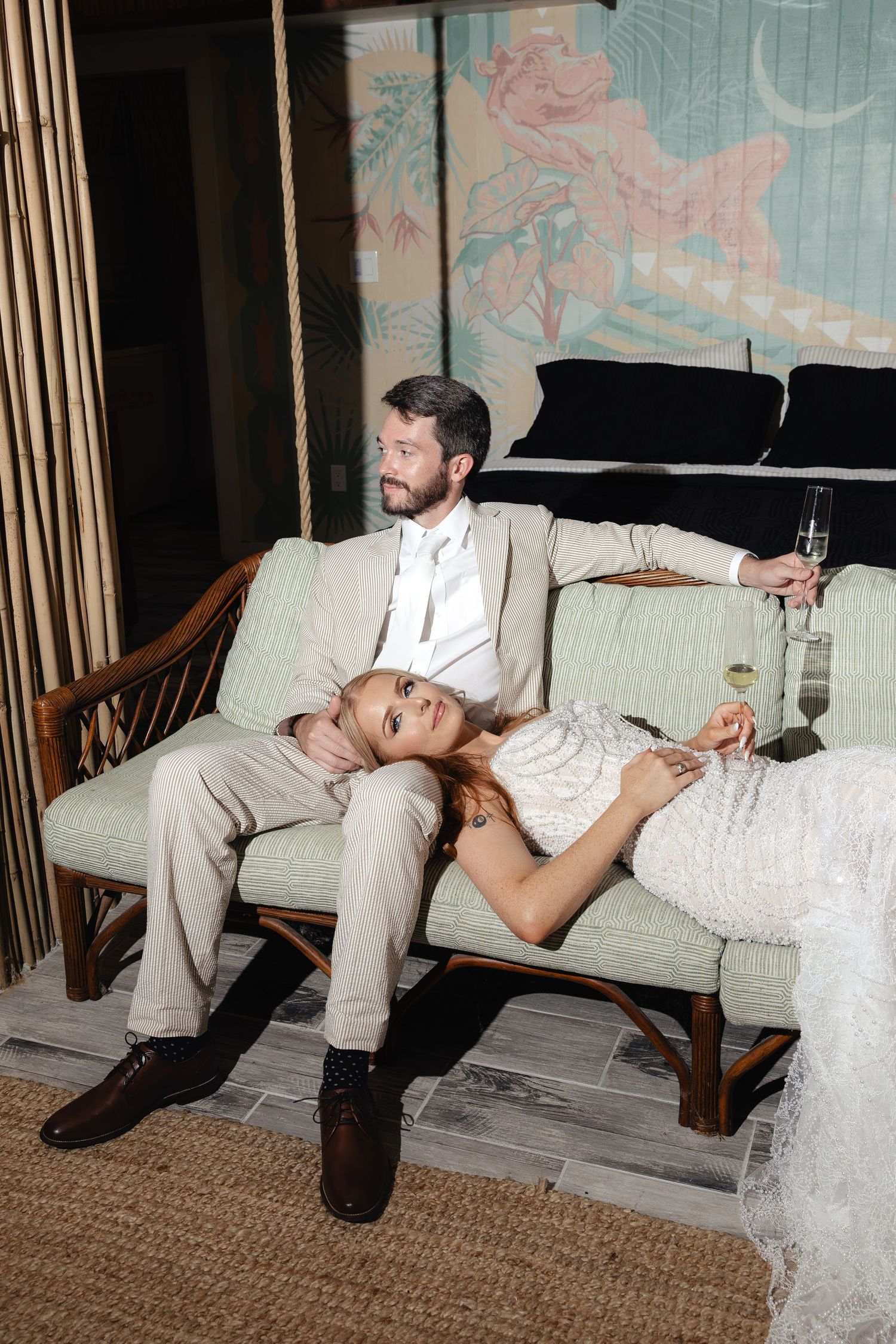 Bride in white lace gown reclines on groom's lap on a rattan sofa, both holding champagne glasses at their wedding reception.