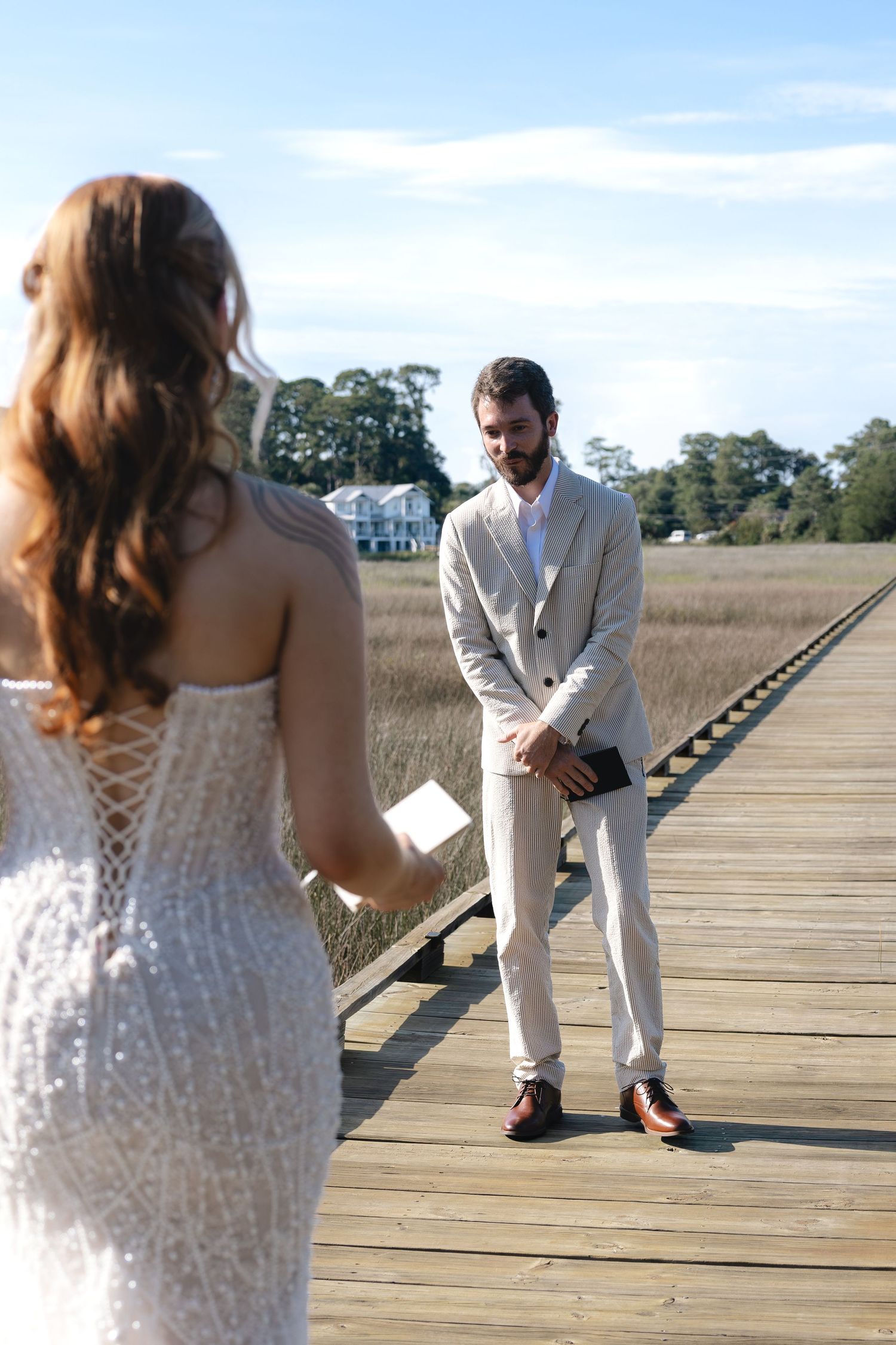 Bride in lace gown reads vows to groom in tan suit on wooden boardwalk surrounded by marsh grasses.