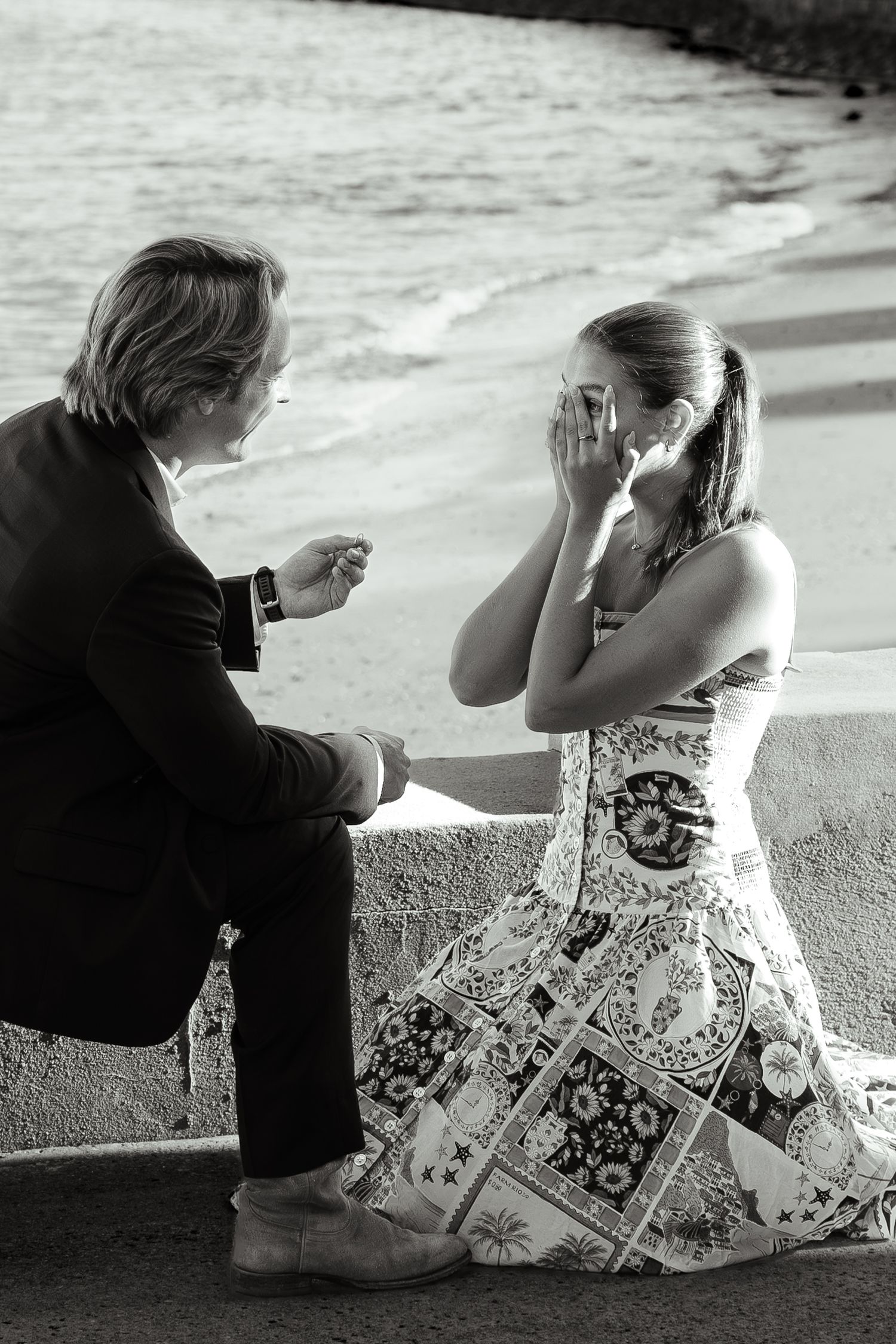 Black and white photo of emotional marriage proposal by the ocean, woman covering her face in joy.