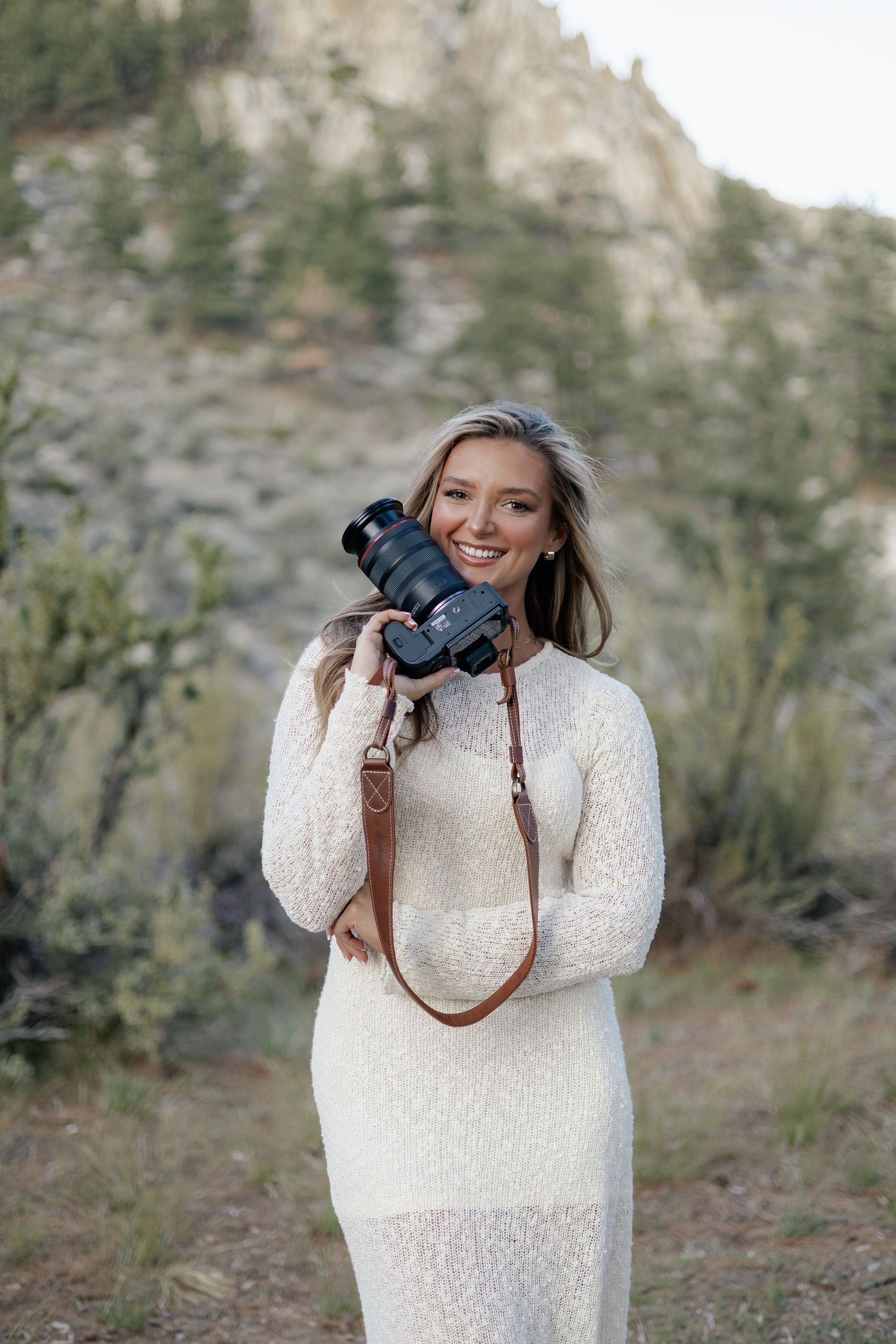 Smiling blonde woman in a white lace dress holding a camera with a brown leather strap in a rocky outdoor setting.