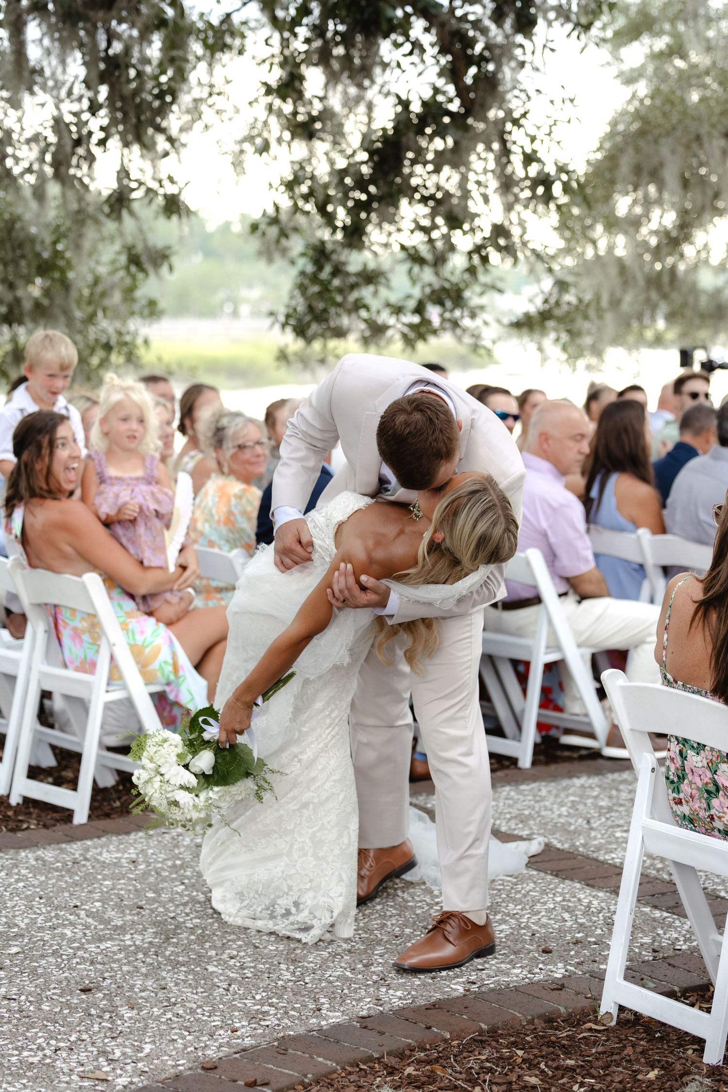 Groom dips bride for a romantic kiss at an outdoor wedding ceremony with guests seated in white chairs.