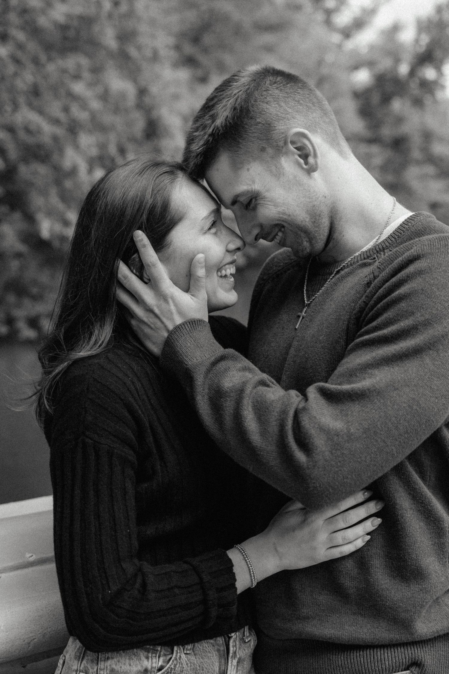 Black and white photo of a laughing couple embracing intimately with foreheads touching near a lake.
