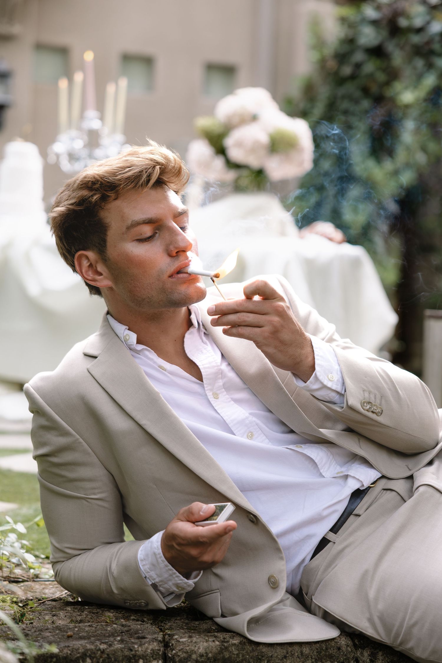 Elegant man in beige suit lighting a cigarette outdoors at a wedding reception.