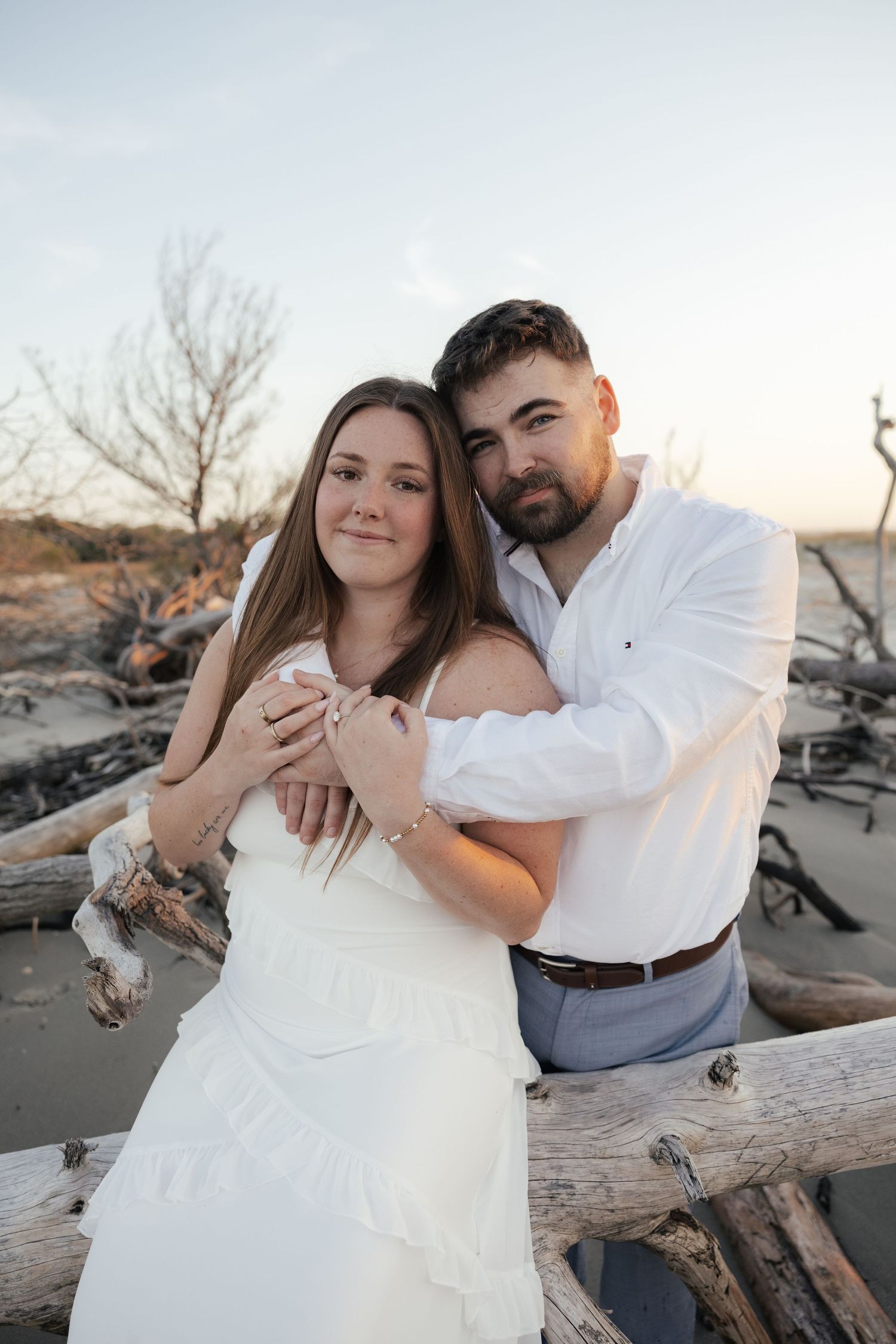 Couple embraces at sunset on driftwood-covered beach, woman in white dress and man in white shirt.