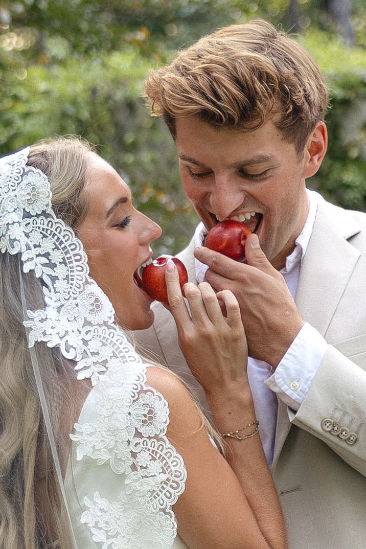 Bride and groom sharing red apples on wedding day, smiling joyfully outdoors in elegant attire.